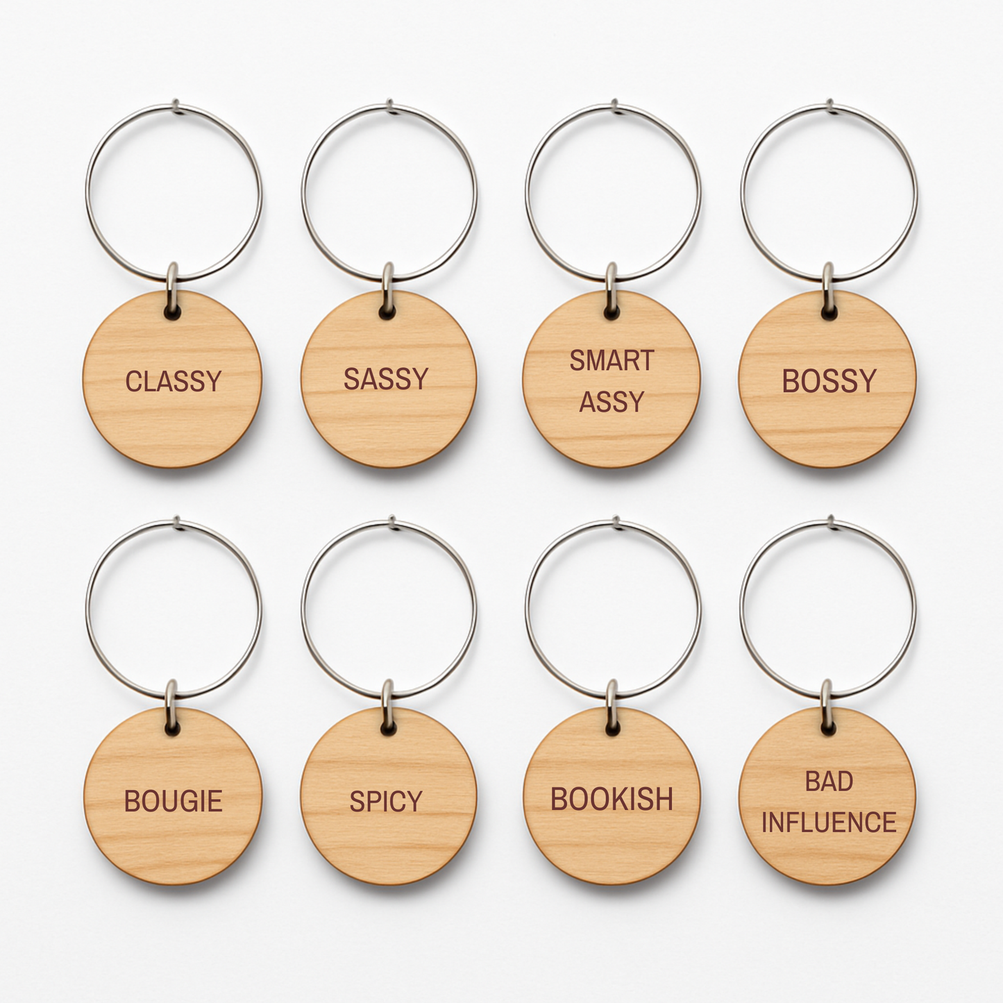 Eight round wooden keychains with metal rings from Mirabilia Boutique’s “Bridal Party Personality Wine Charms | Set of 8” are arranged in two rows. Each charm showcases a distinct engraved word—“CLASSY,” “SASSY,” “SMART ASSY,” “BOSSY,” “BOUGIE,” “SPICY,” “BOOKISH,” and “BAD INFLUENCE.” Perfect as unique bridal party wine charms or thoughtful gifts, these wooden accents add a playful touch to any celebration.