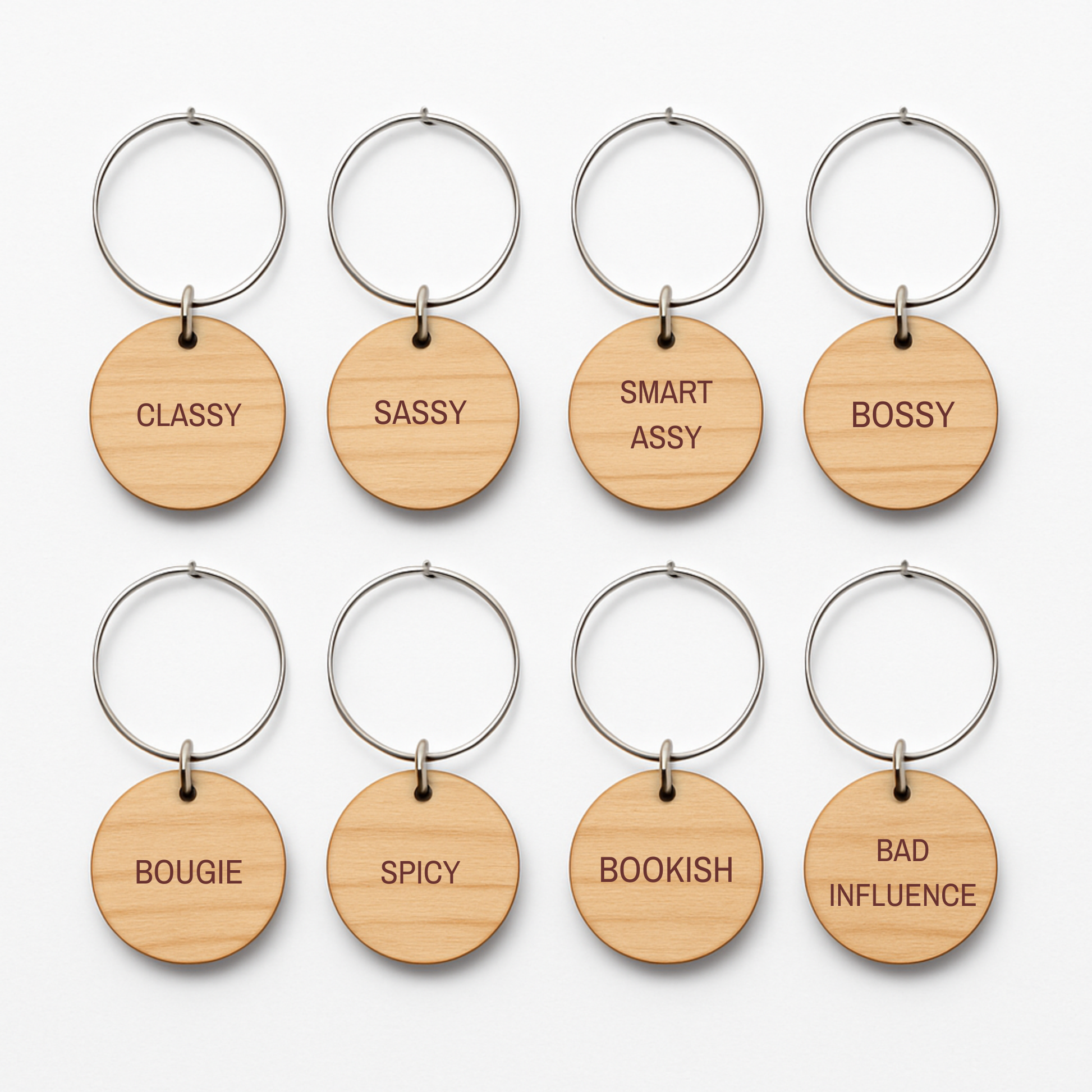 Eight round wooden keychains with metal rings from Mirabilia Boutique’s “Bridal Party Personality Wine Charms | Set of 8” are arranged in two rows. Each charm showcases a distinct engraved word—“CLASSY,” “SASSY,” “SMART ASSY,” “BOSSY,” “BOUGIE,” “SPICY,” “BOOKISH,” and “BAD INFLUENCE.” Perfect as unique bridal party wine charms or thoughtful gifts, these wooden accents add a playful touch to any celebration.