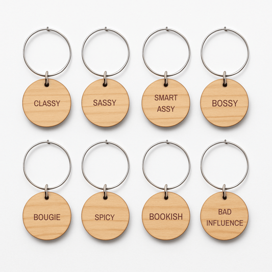 Eight round wooden keychains with metal rings from Mirabilia Boutique’s “Bridal Party Personality Wine Charms | Set of 8” are arranged in two rows. Each charm showcases a distinct engraved word—“CLASSY,” “SASSY,” “SMART ASSY,” “BOSSY,” “BOUGIE,” “SPICY,” “BOOKISH,” and “BAD INFLUENCE.” Perfect as unique bridal party wine charms or thoughtful gifts, these wooden accents add a playful touch to any celebration.