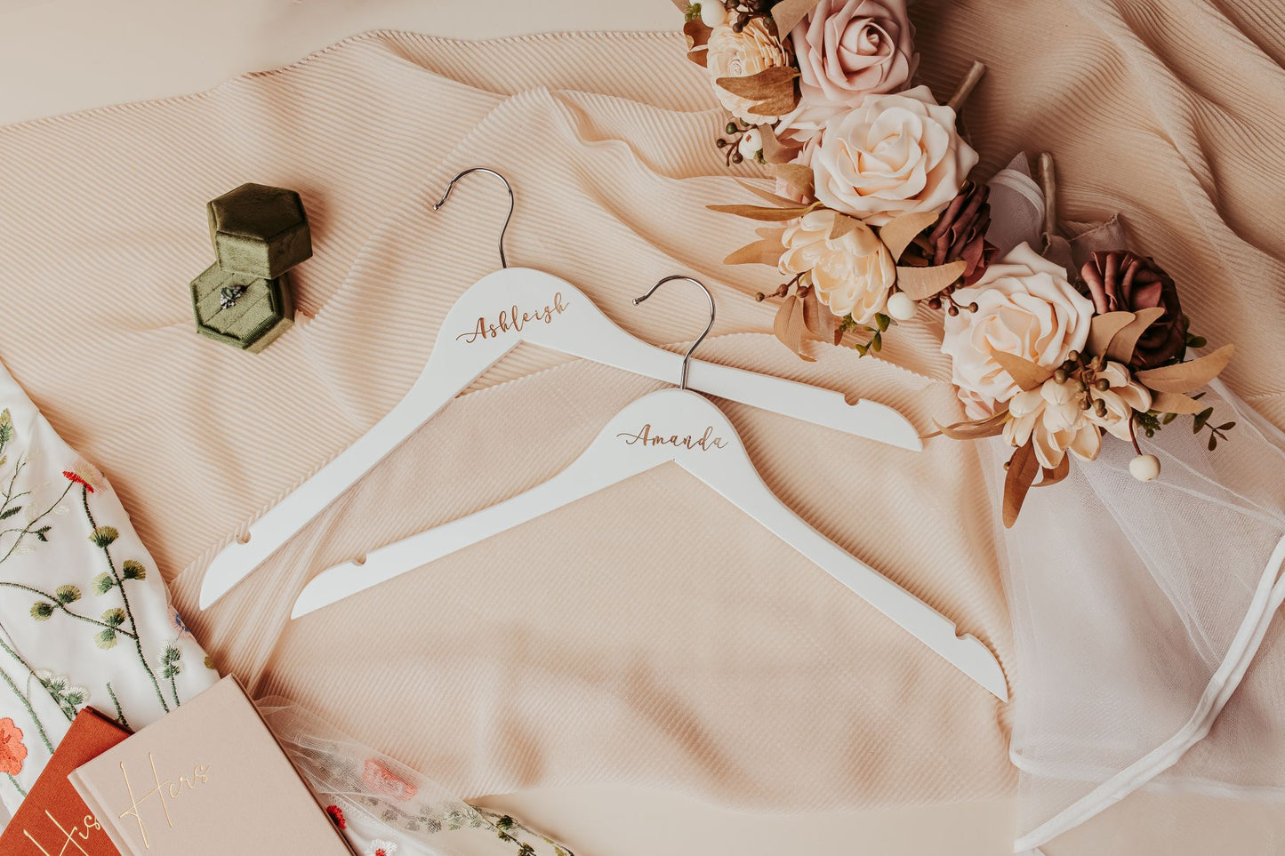 The Mirabilia Boutique Personalized Wedding Hangers | Custom Name Hanger Set, featuring names in elegant script, rest on beige fabric with flowers and a green ring box, adding a soft, romantic touch to your wedding day details.