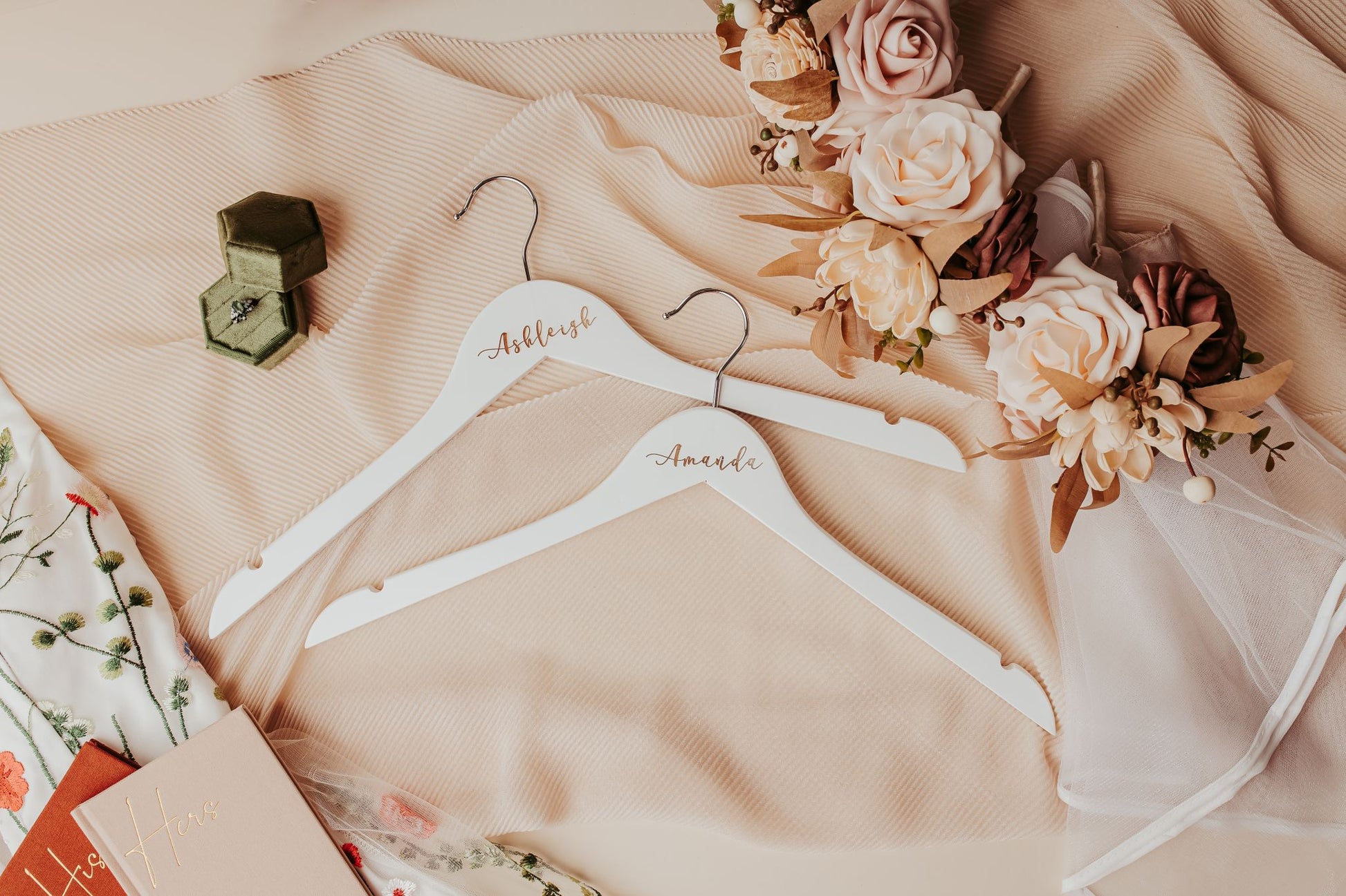 The Mirabilia Boutique Personalized Wedding Hangers | Custom Name Hanger Set, featuring names in elegant script, rest on beige fabric with flowers and a green ring box, adding a soft, romantic touch to your wedding day details.