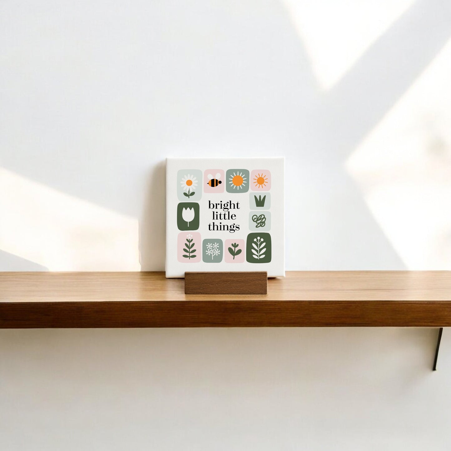 The Bright Little Things Mini Canvas Sign by Mirabilia Boutique features simple flower illustrations and an uplifting phrase, adding handcrafted charm to a wooden shelf as soft natural light casts gentle shadows on the white wall.