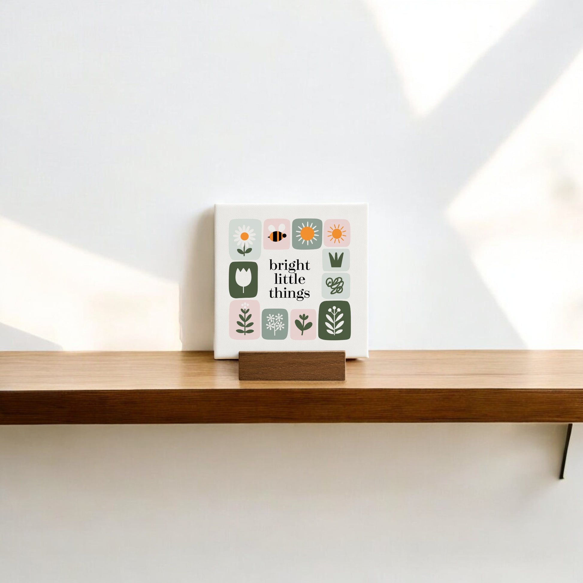 The Bright Little Things Mini Canvas Sign by Mirabilia Boutique features simple flower illustrations and an uplifting phrase, adding handcrafted charm to a wooden shelf as soft natural light casts gentle shadows on the white wall.
