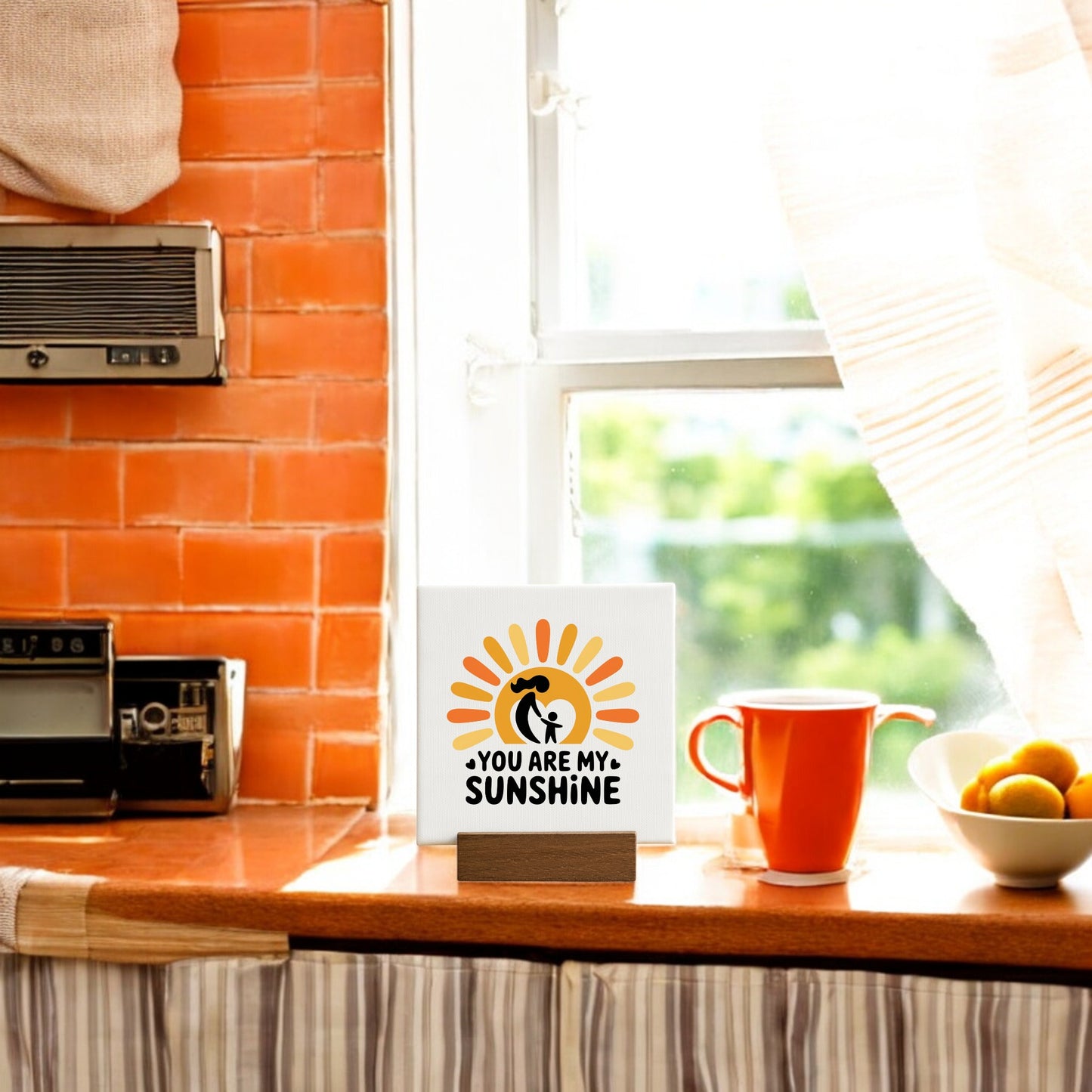 A kitchen windowsill displays the Mirabilia Boutique You Are My Sunshine Mini Sign – Mom & Child Canvas beside an orange mug and a bowl of oranges. Sunlight highlights this cozy, meaningful decor—perfect for Mother's Day gifting.