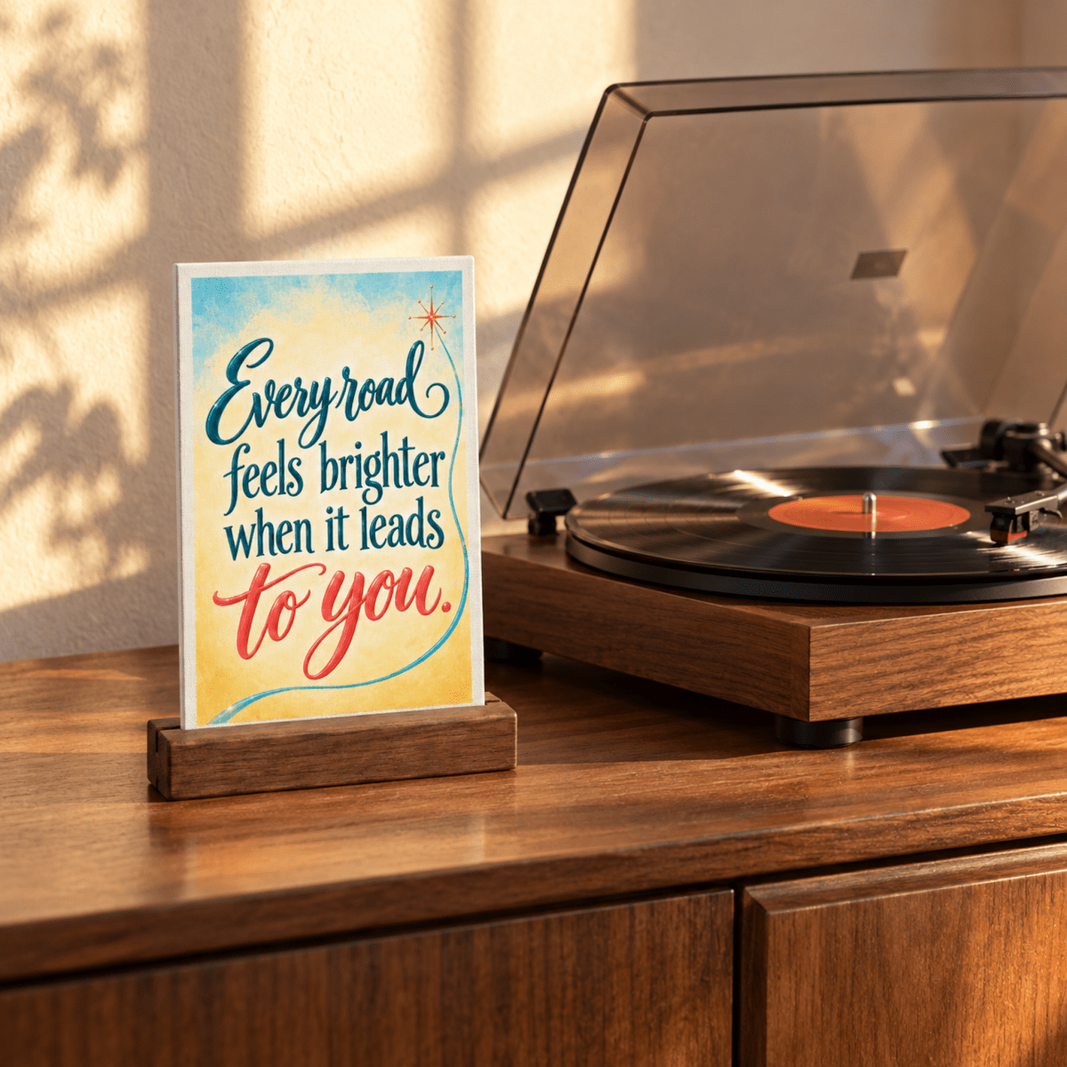 The Every Road Feels Brighter Mini Sign | Chrome & Starlight sits in a wood stand on a cabinet next to a record player, displaying the expressive lettering “Every road feels brighter when it leads to you.”