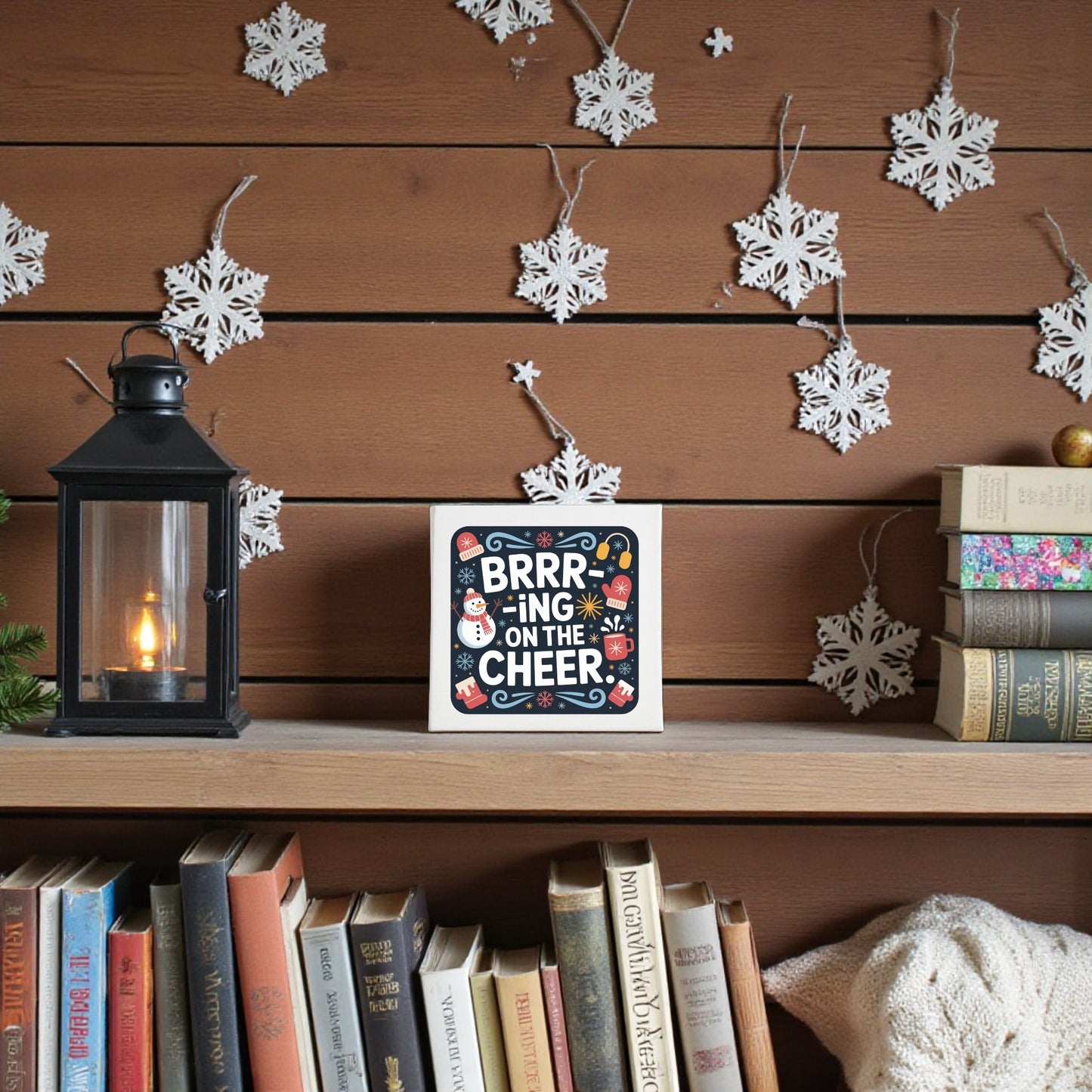 A cozy wooden shelf shows the “Brrr-ing on the Cheer Canvas | 8x8 Holiday Wall Sign” by Mirabilia Boutique, a lantern, and books. White paper snowflakes on wood-paneled walls add festive charm to your holiday home decor.