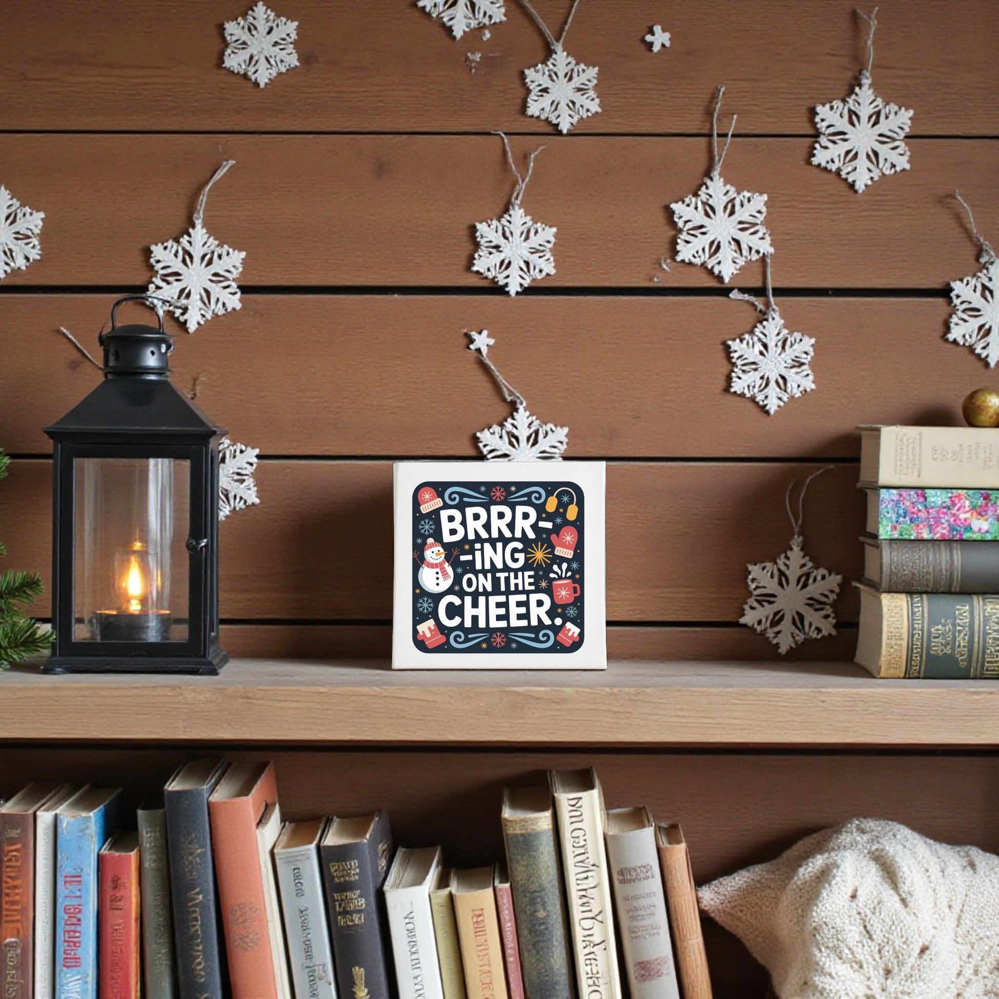 A cozy wooden shelf shows the “Brrr-ing on the Cheer Canvas | 8x8 Holiday Wall Sign” by Mirabilia Boutique, a lantern, and books. White paper snowflakes on wood-paneled walls add festive charm to your holiday home decor.