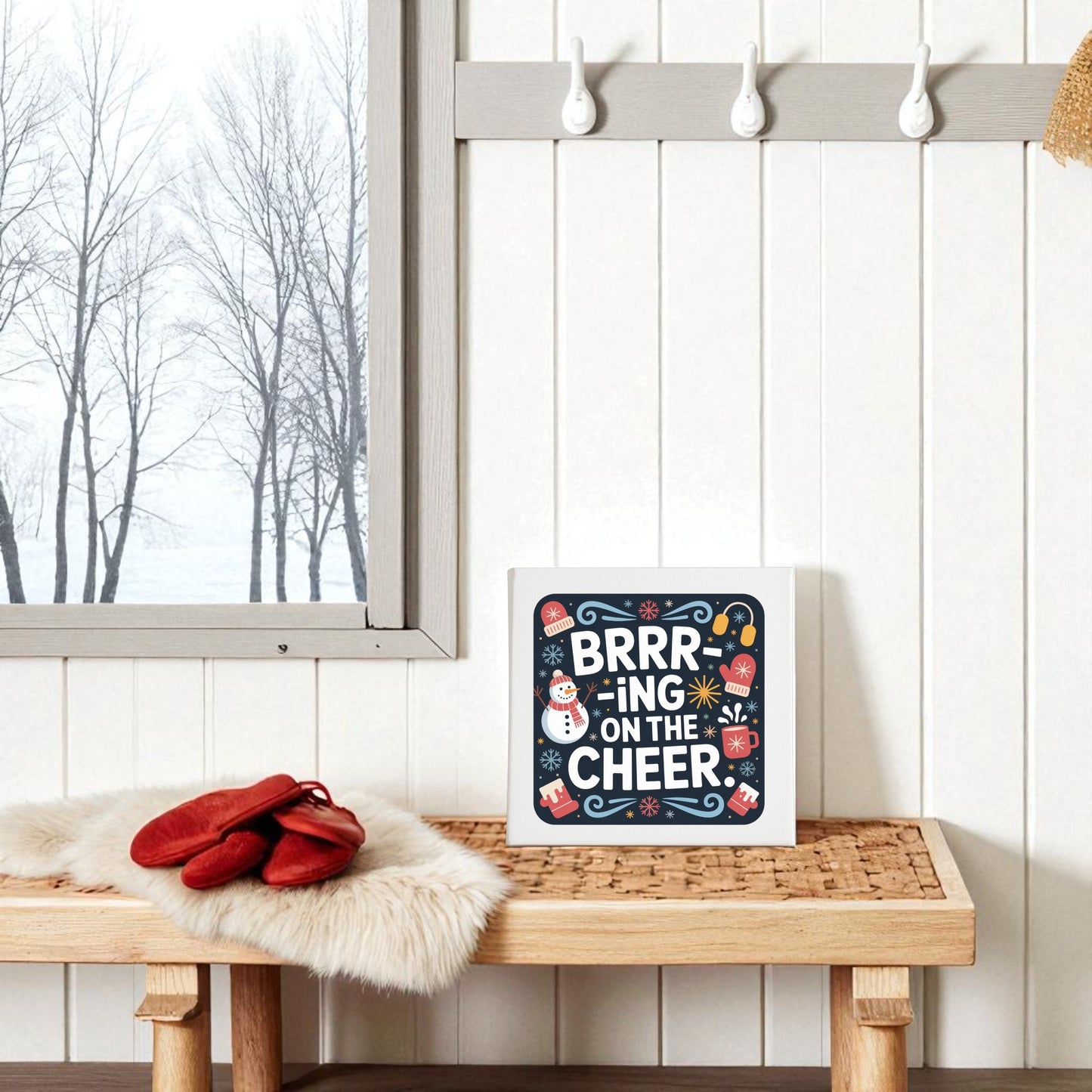 A cozy winter entryway features Mirabilia Boutique's "Brrr-ing on the Cheer Canvas | 8x8 Holiday Wall Sign," red mittens on a fur mat, and snowy trees by a frosted window—creating the perfect festive home decor backdrop.