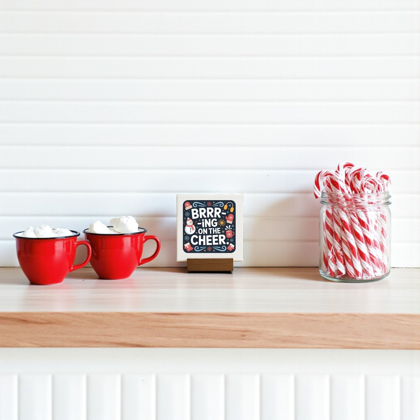 Two red mugs with marshmallows, a Mirabilia Boutique Mini Brrr-ing on the Cheer Sign (4x4), and a glass jar of candy canes make festive holiday tiered tray decor on a wooden shelf against a white background.