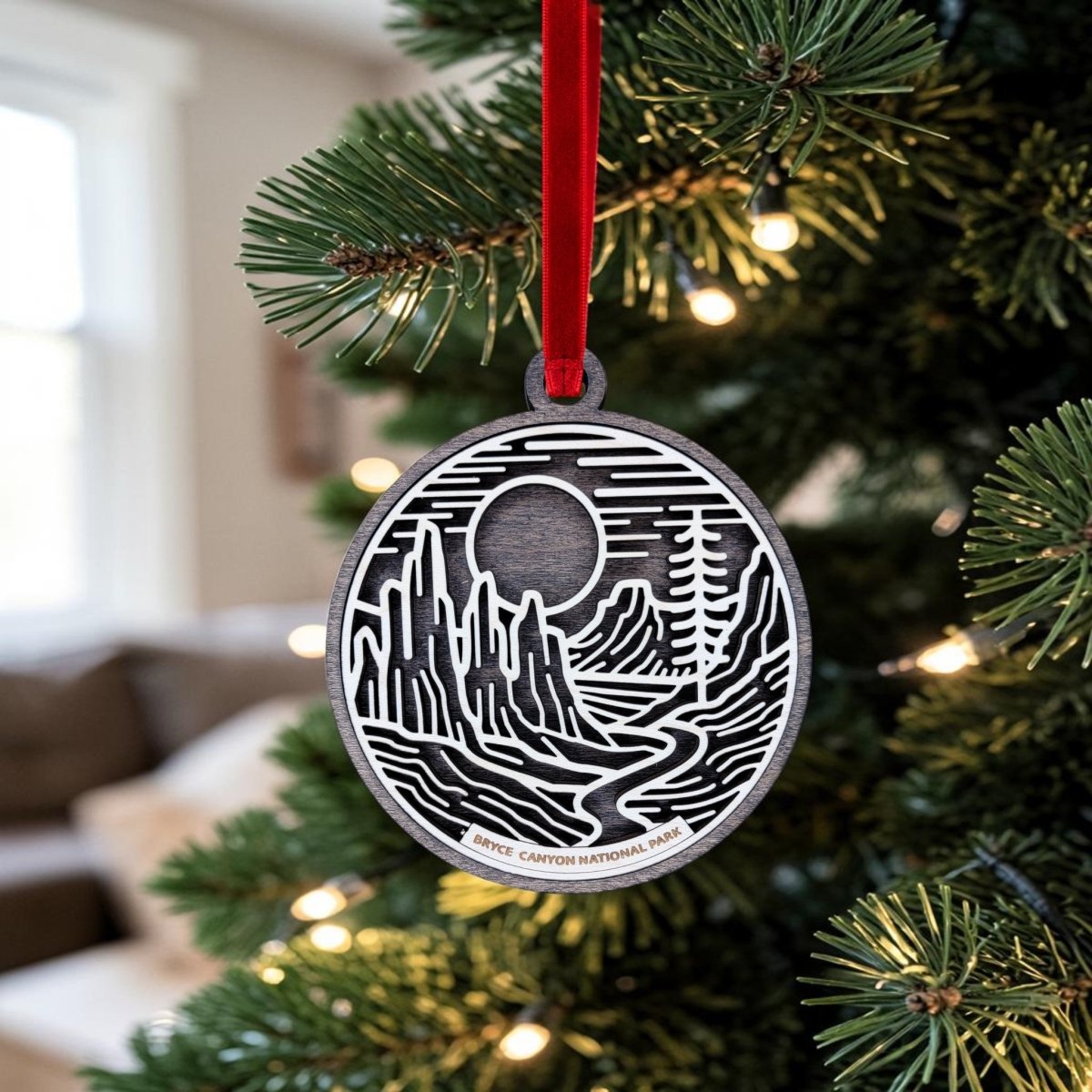 A round wooden Bryce Canyon National Park Christmas Ornament by Mirabilia Boutique, featuring black and white line art of mountains, a river, and the words "Bryce Canyon National Park," hangs on a Christmas tree with a red ribbon.