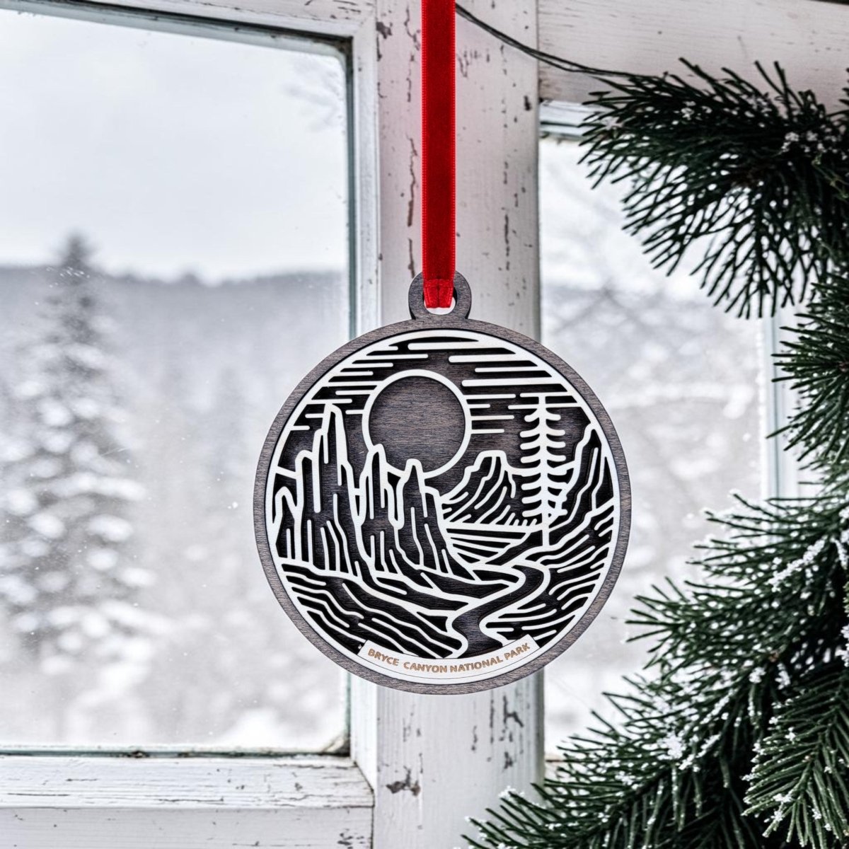 A Bryce Canyon National Park Christmas Ornament from Mirabilia Boutique, featuring round wooden line art with a landscape design, hangs by a red ribbon in front of a snowy window adorned with pine branches. Outside, snow-covered trees evoke the serene beauty of the park.