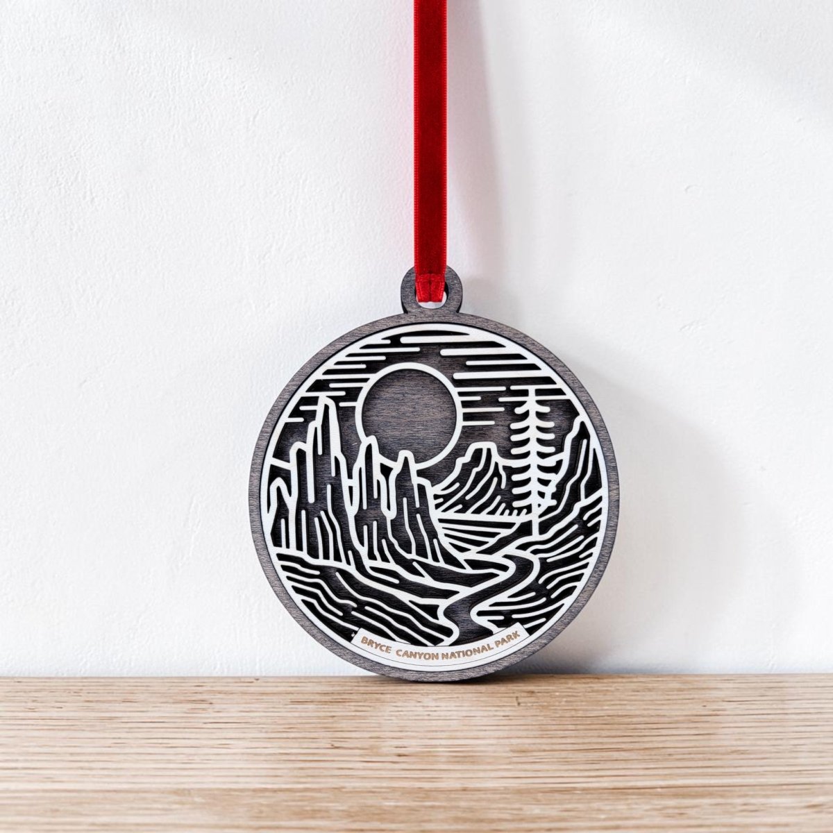The Mirabilia Boutique Bryce Canyon National Park Christmas Ornament is a round wooden ornament featuring a black and white line art illustration of Bryce Canyon’s striking rock formations beneath a sun or moon. It hangs by a red ribbon and is shown resting against a white wall on a wooden surface.