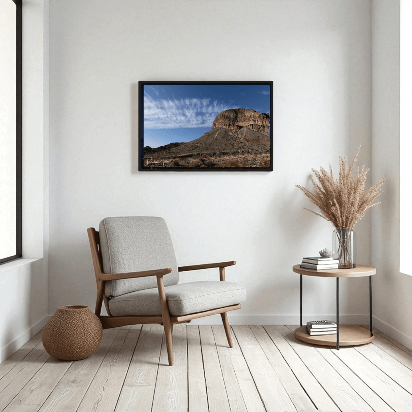 A minimalist living room featuring a beige armchair, round wooden side table with dried grasses, woven basket, and the Burro Mesa Pouroff Print | Big Bend Framed Wall Art by Mirabilia Boutique. Light wood floors and neutral decor complete the look.