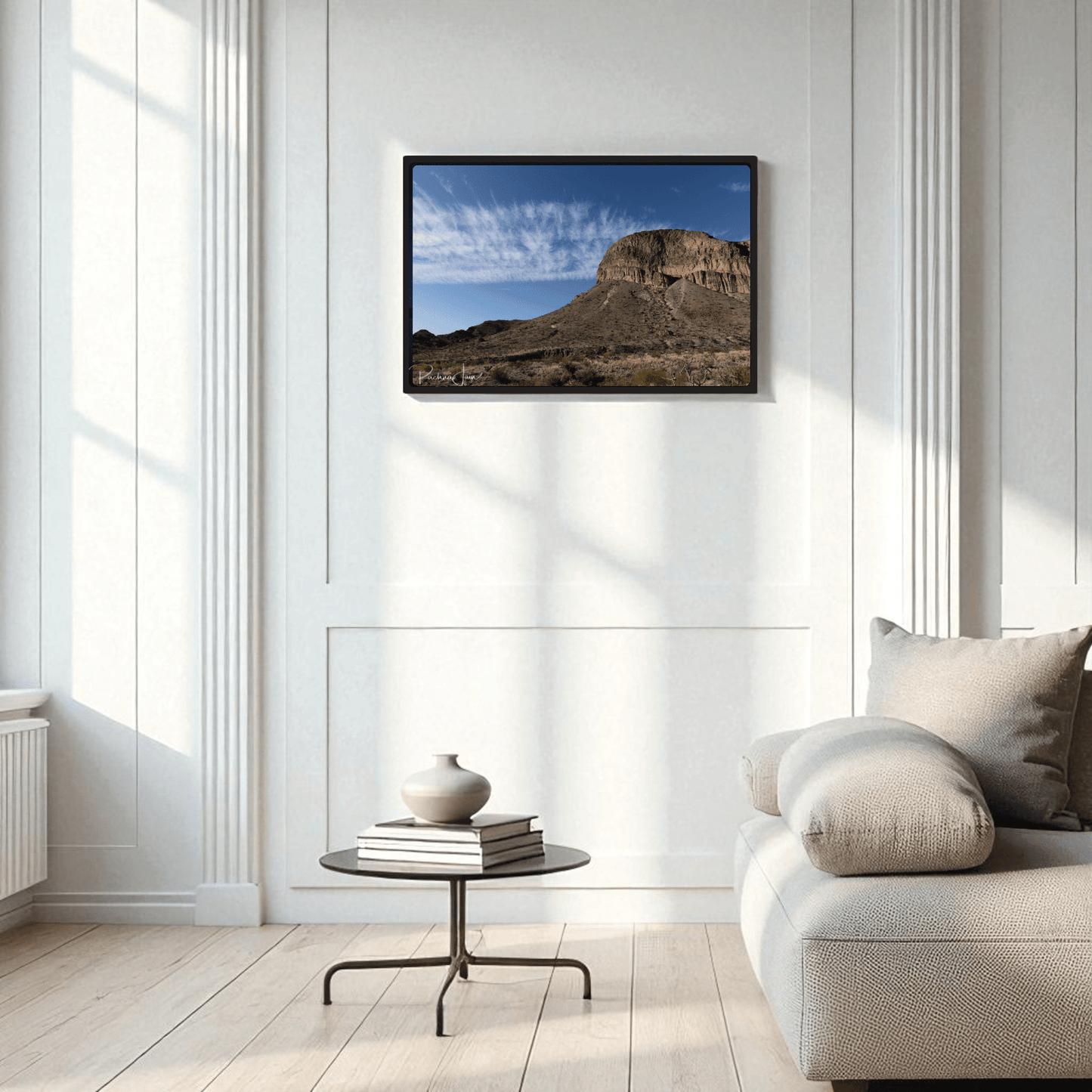 A minimalist living room with a light gray sofa, round black coffee table, and vase features the Burro Mesa Pouroff Print | Big Bend Framed Wall Art by Mirabilia Boutique on a white paneled wall.