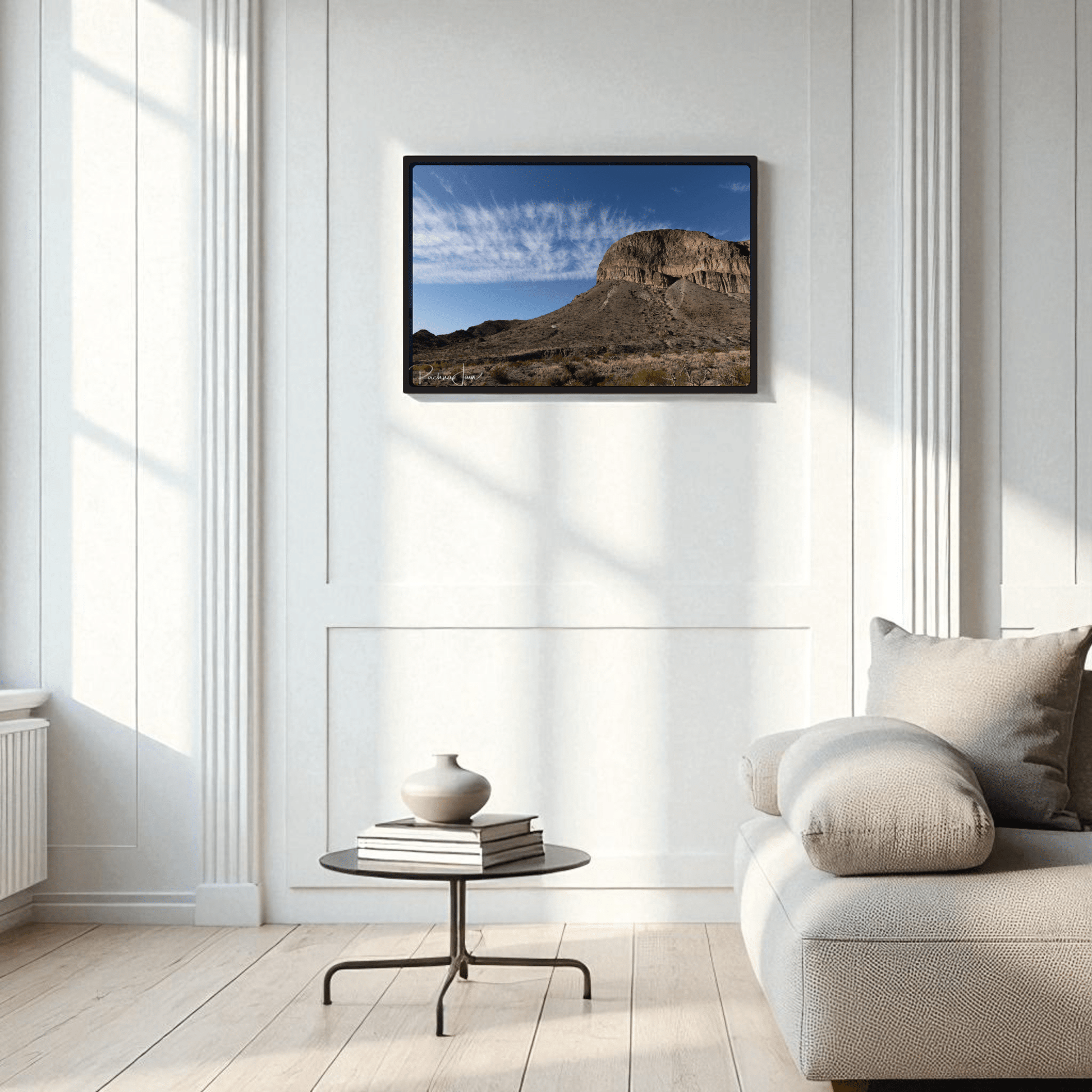 A minimalist living room with a light gray sofa, round black coffee table, and vase features the Burro Mesa Pouroff Print | Big Bend Framed Wall Art by Mirabilia Boutique on a white paneled wall.