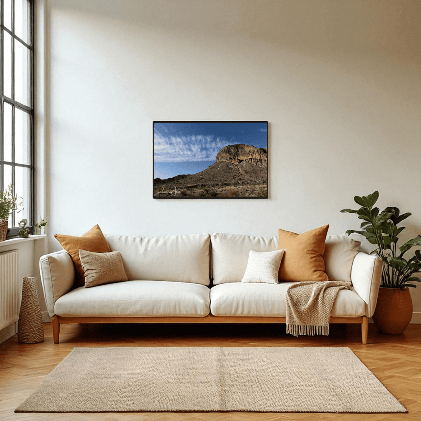 A modern living room features a white sofa with tan and brown pillows, a beige throw, houseplants, and Mirabilia Boutique’s Burro Mesa Pouroff Print | Big Bend Framed Wall Art above the sofa. Sunlight pours in through large windows.