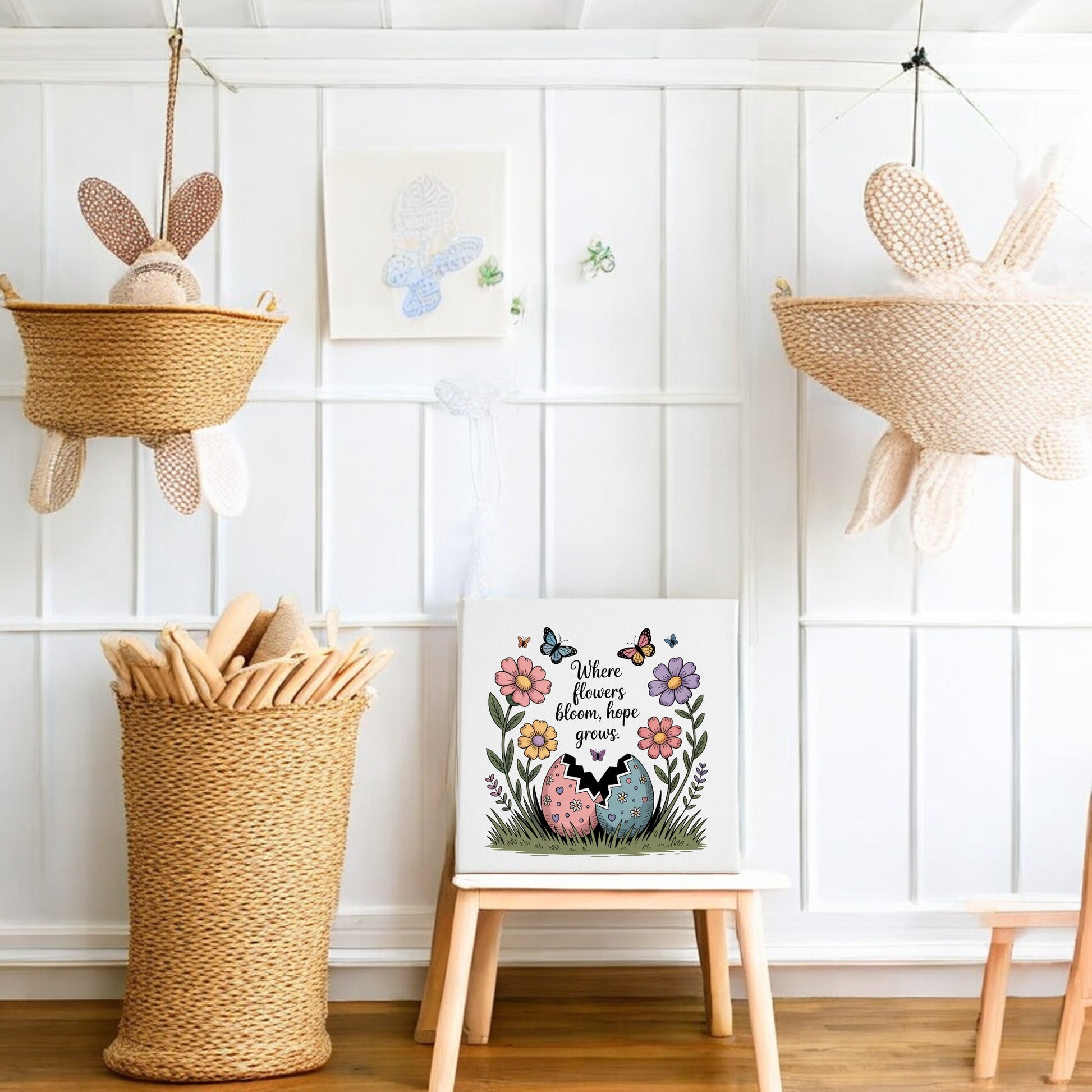 A cozy, white-walled nursery with spring decor features two woven baskets with stuffed bunnies, a wicker basket of wooden toys, and the Mirabilia Boutique "Where Flowers Bloom, Hope Grows" 8x8 Spring Canvas, accented by colorful floral wall art.