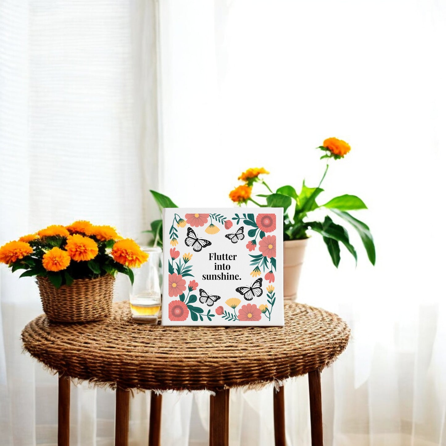 A round wicker table displays orange flowers, a juice glass, and the Mirabilia Boutique “Flutter into Sunshine Canvas – Butterflies & Floral Joy.” Sheer curtains and natural light enhance the cozy space with inspirational wall art.