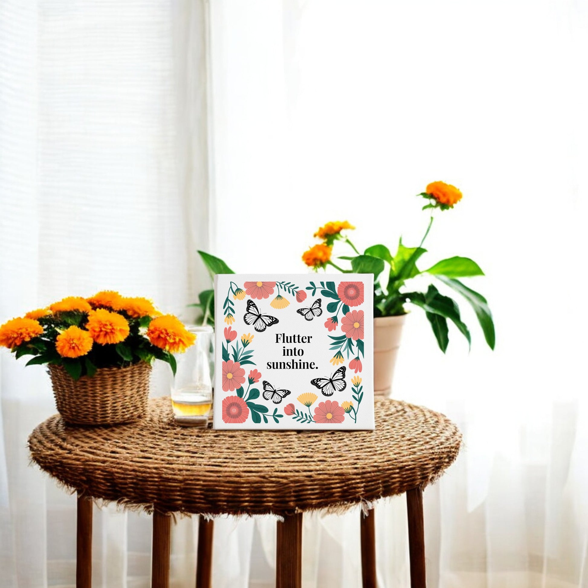 A round wicker table displays orange flowers, a juice glass, and the Mirabilia Boutique “Flutter into Sunshine Canvas – Butterflies & Floral Joy.” Sheer curtains and natural light enhance the cozy space with inspirational wall art.