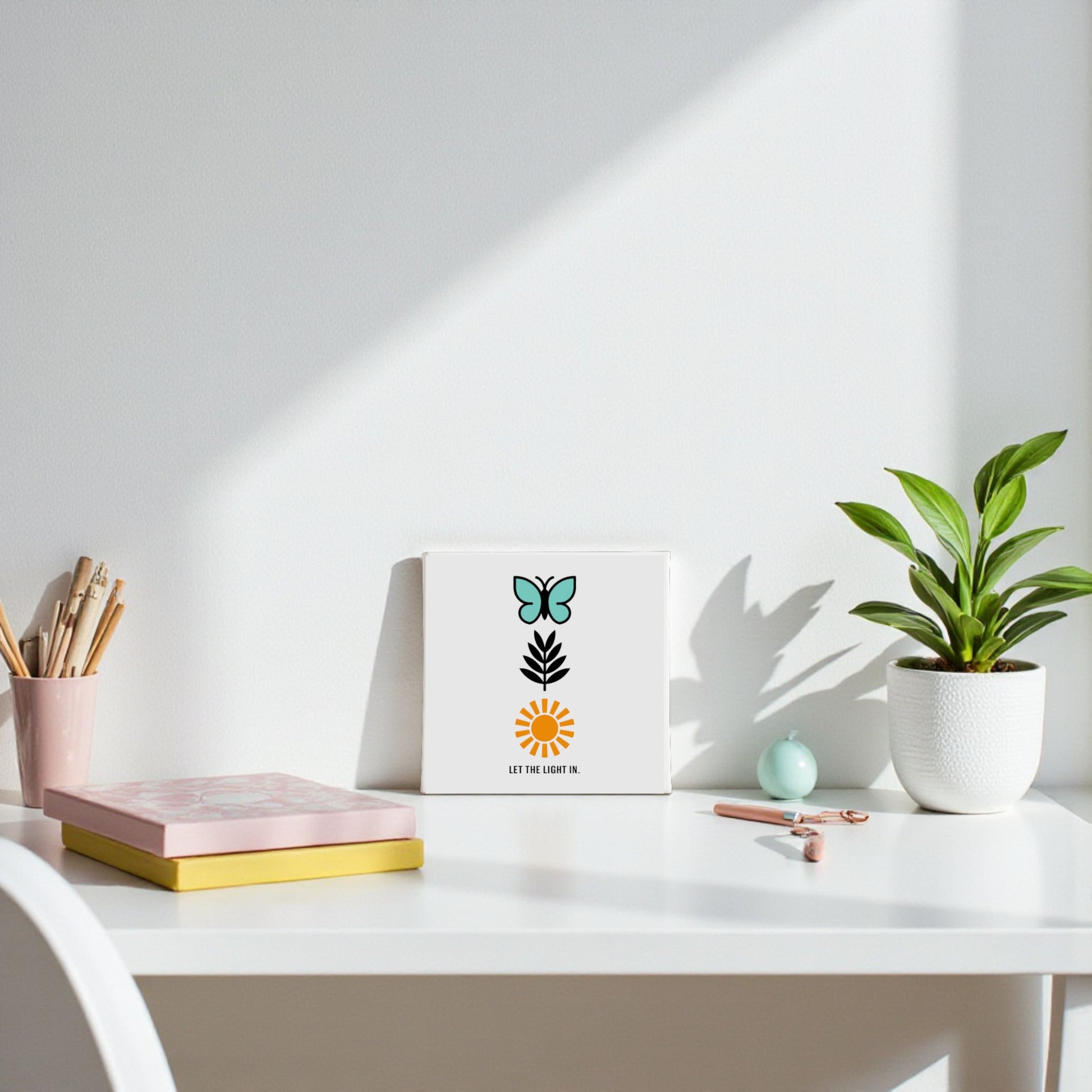 A minimal white desk with a plant, notebooks, pencils, and the "Let the Light In Canvas – Butterfly Sun Wall Art" by Mirabilia Boutique, featuring inspiring decor and the words "LET THE LIGHT IN," all illuminated by natural sunlight.