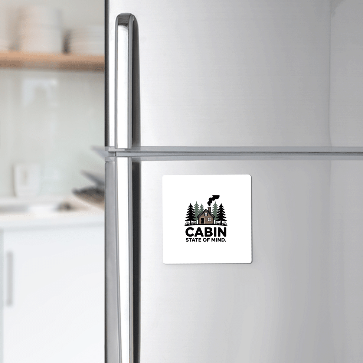 A Mirabilia Boutique "Cabin State of Mind Magnet" with a cozy cabin and trees design adds rustic charm to a stainless steel refrigerator, while dishes sit on the kitchen counter in the background.
