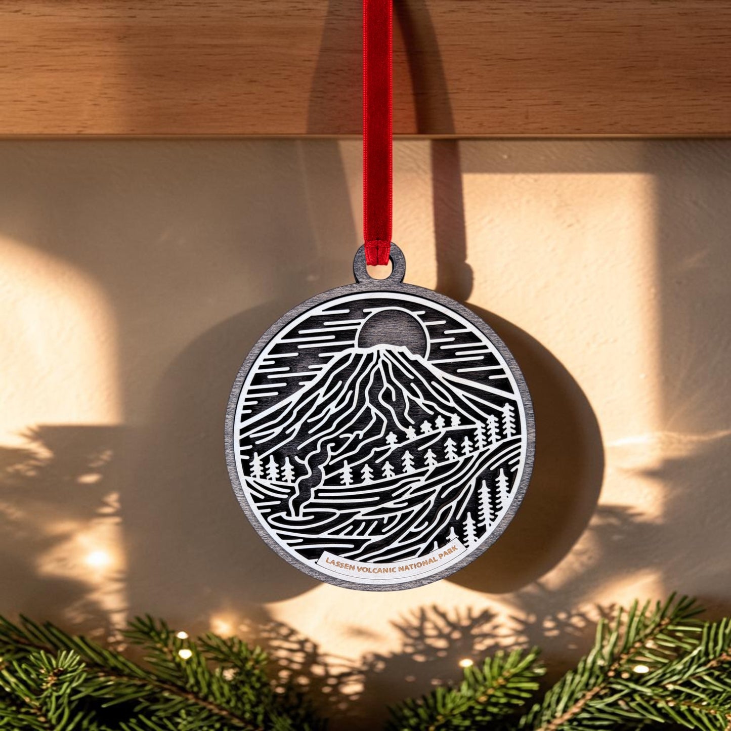 The Lassen Volcanic National Park Line Art Christmas Ornament by Mirabilia Boutique features a round wooden design with a striking black and white illustration of a mountain, trees, and sun, capturing the spirit of Lassen Peak. It hangs from a red ribbon, with "Lassen Volcanic National Park" inscribed below. Pine branches are pictured at the bottom.