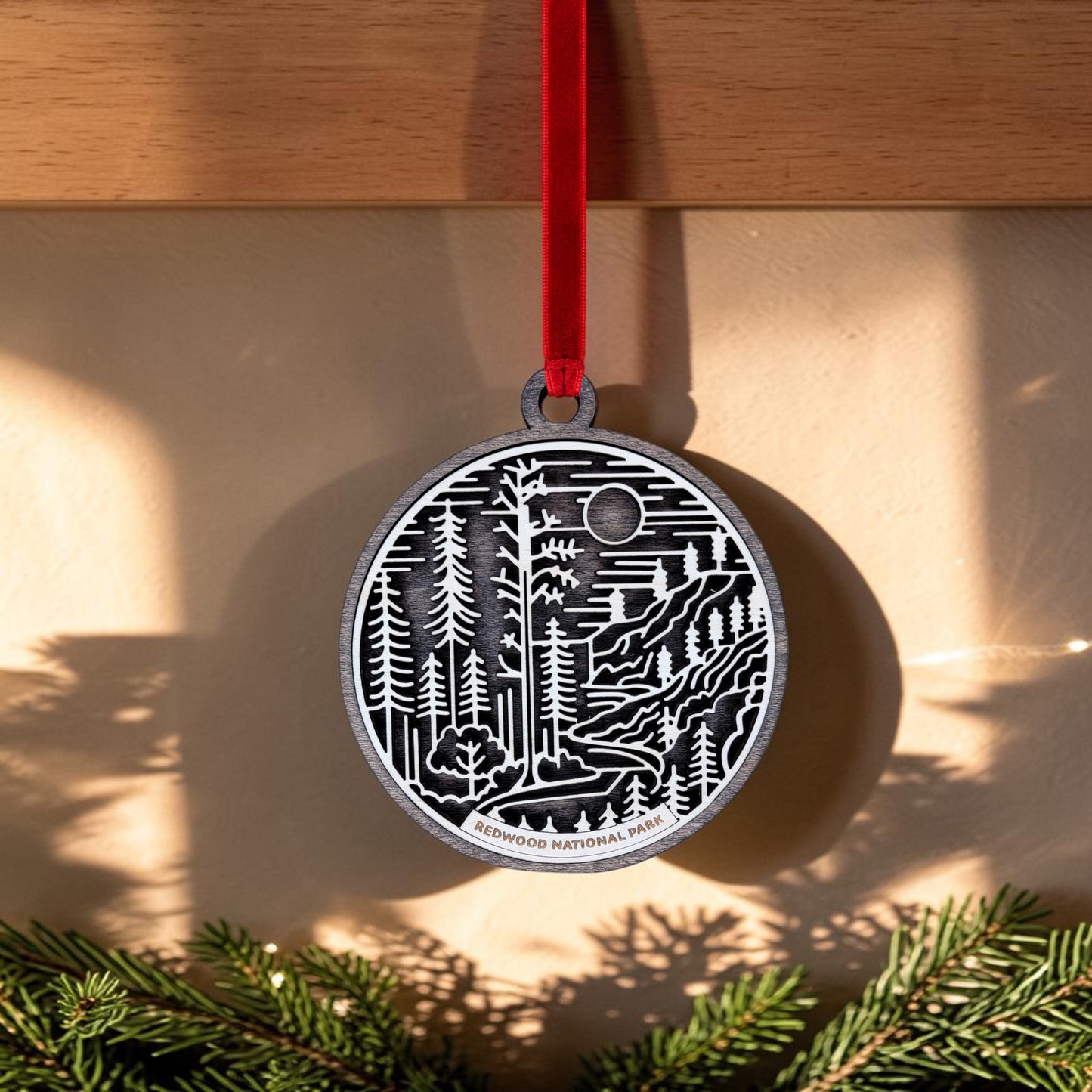 A handcrafted Redwood National Park Line Art Christmas Ornament by Mirabilia Boutique, featuring black and white layered forest artwork and the words "Redwood National Park," hangs from a red ribbon, casting a shadow on a beige wall above green pine branches. The detailed line art highlights the natural beauty of this distinctive ornament.