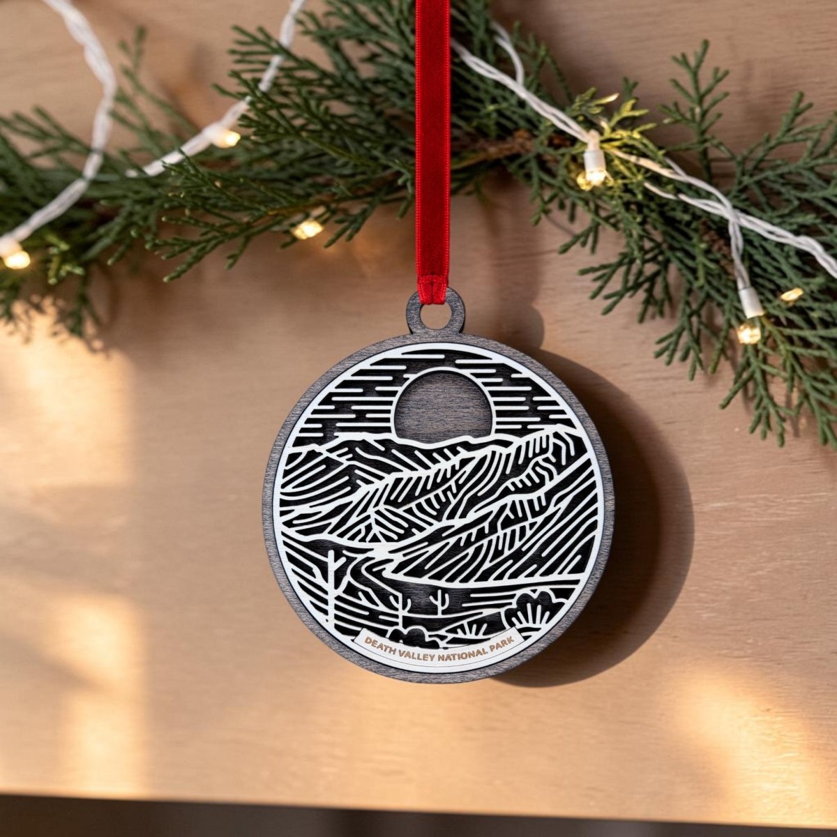 The Death Valley National Park Christmas Ornament by Mirabilia Boutique features a handcrafted wooden oval design with line art of the desert landscape and "Death Valley National Park" text. Hanging from a red ribbon, this unique keepsake is displayed in front of greenery and string lights.