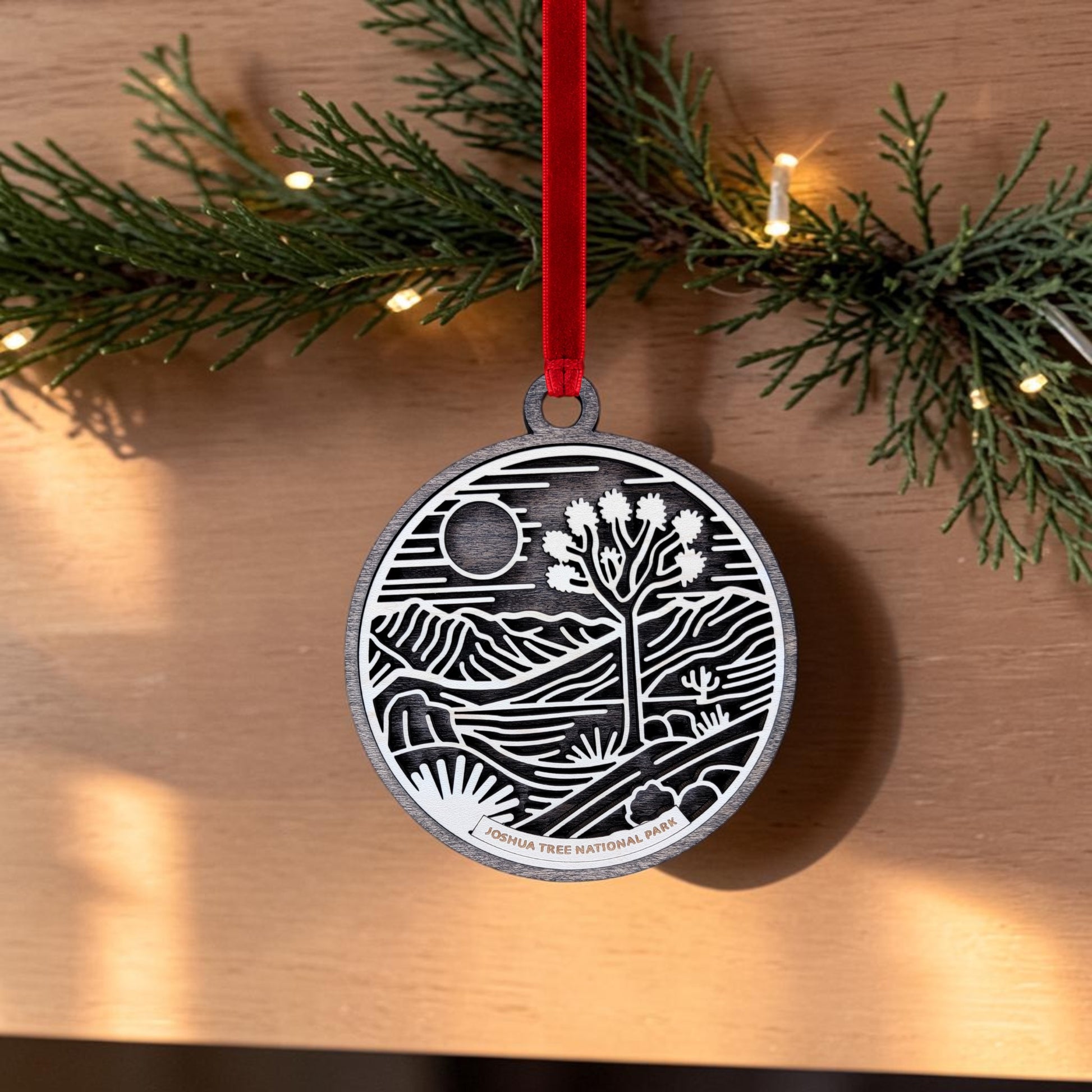 The Joshua Tree National Park Line Art Christmas Ornament by Mirabilia Boutique is an oval wooden ornament engraved with a desert landscape, including a Joshua tree, mountains, and the sun. It hangs from greenery by a red ribbon, with string lights visible in the background.