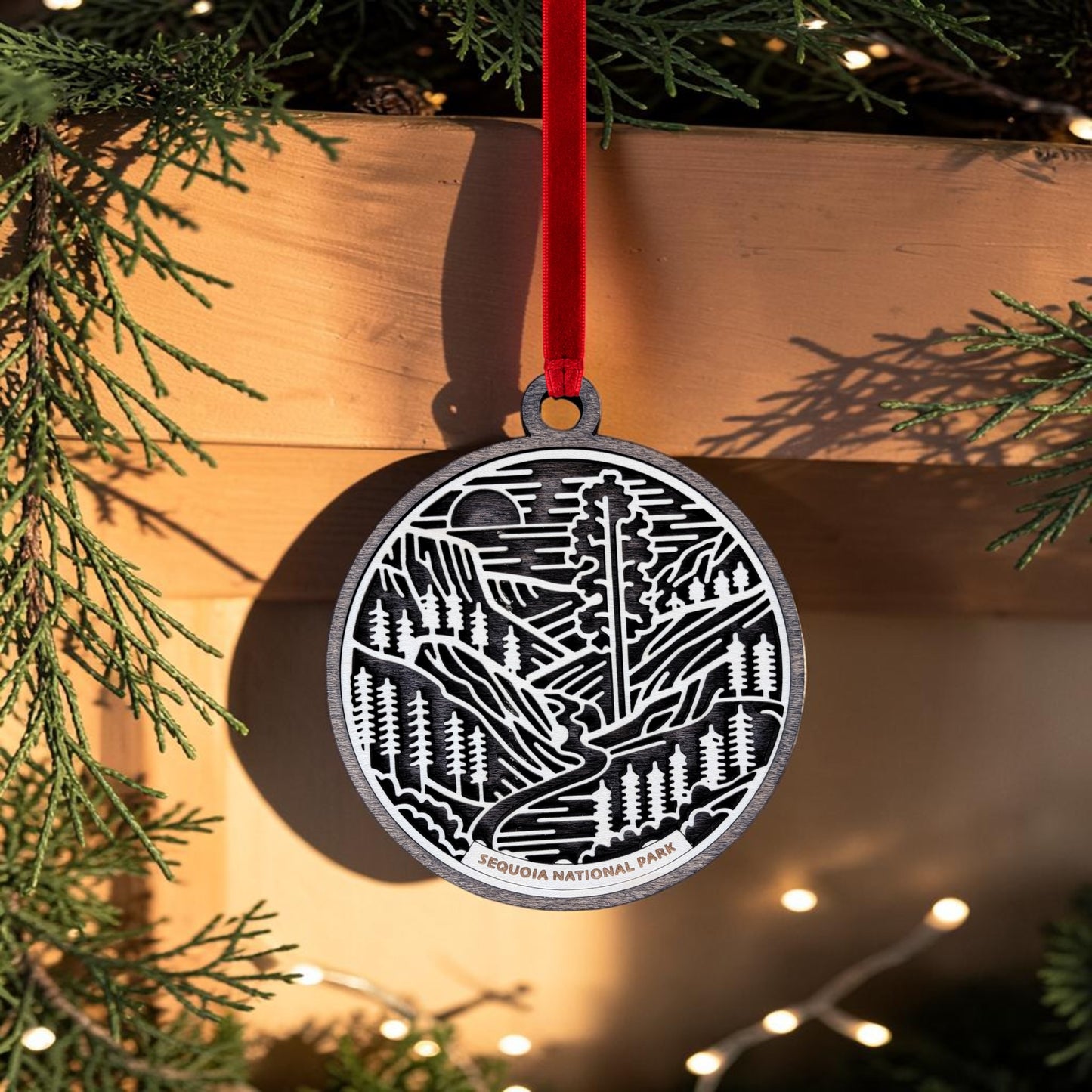 The Sequoia National Park Line Art Christmas Ornament by Mirabilia Boutique features an elegant wooden design with a Sequoia tree engraving, hanging from a red ribbon amid twinkling lights and lush pine branches—a perfect giant sequoia keepsake for your holiday collection.