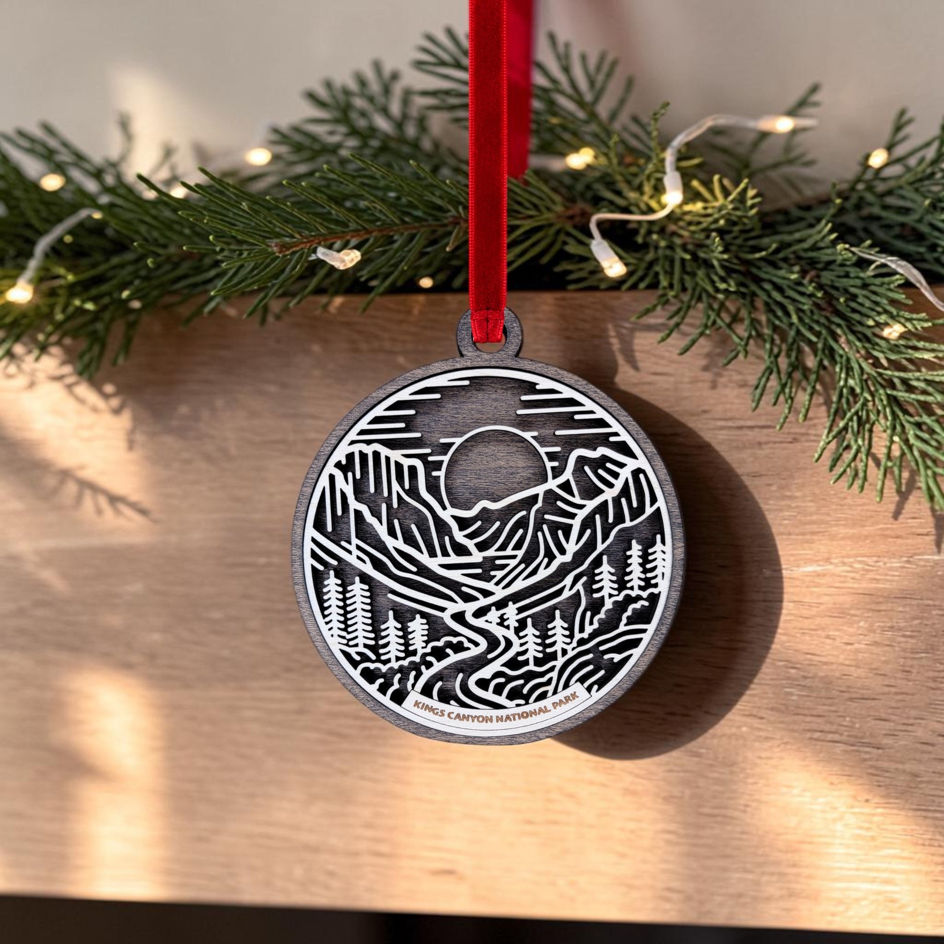 The Kings Canyon National Park Line Art Christmas Ornament by Mirabilia Boutique features a metal design with a mountain and forest scene, suspended from a red ribbon. Displayed in front of greenery and string lights on a wooden surface, the ornament celebrates the majesty of giant sequoias with the inscription "Kings Canyon National Park.