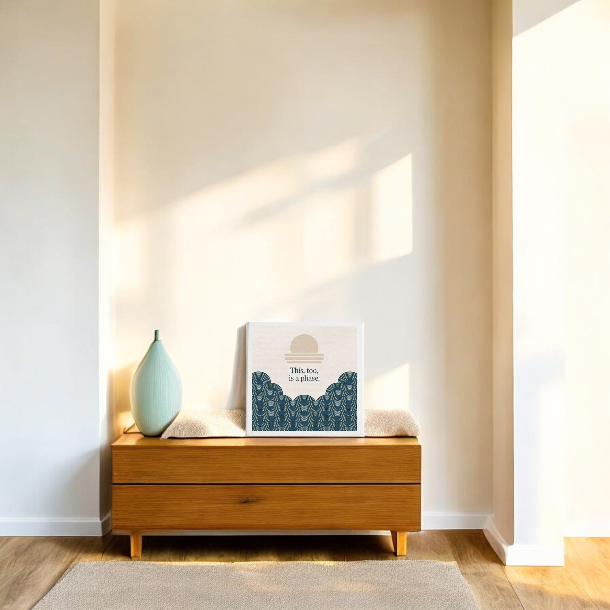 A wooden bench with a soft cushion sits by a sunlit wall. Atop it, a teal vase stands next to framed abstract seascape art and Mirabilia Boutique's "This Too Is a Phase Canvas – Calming Summer Wall Art," bathed in natural light.