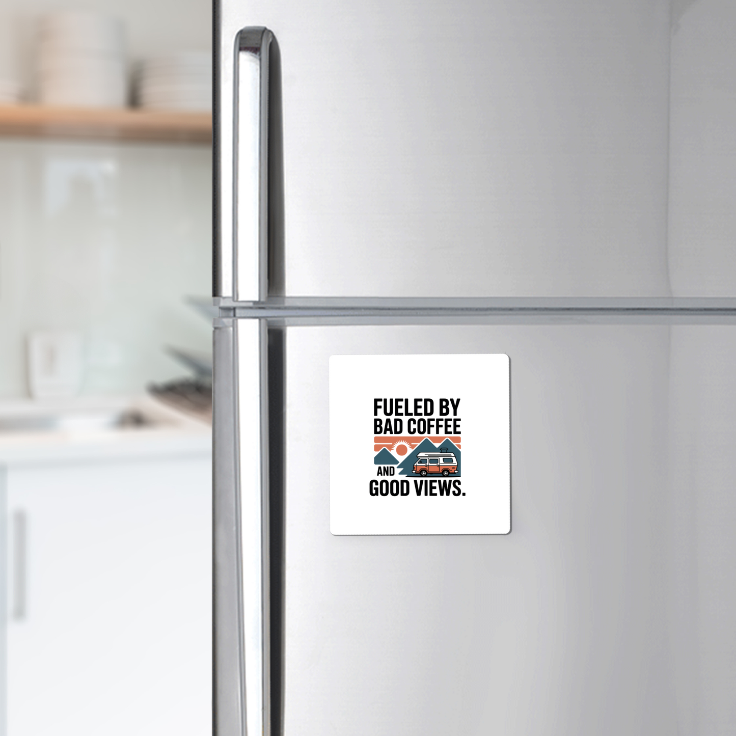 A silver refrigerator displays the "Fueled by Bad Coffee and Good Views Magnet" from Mirabilia Boutique—a fun camping souvenir with a coffee mug and mountain graphic—above a kitchen counter with dishes in the background.
