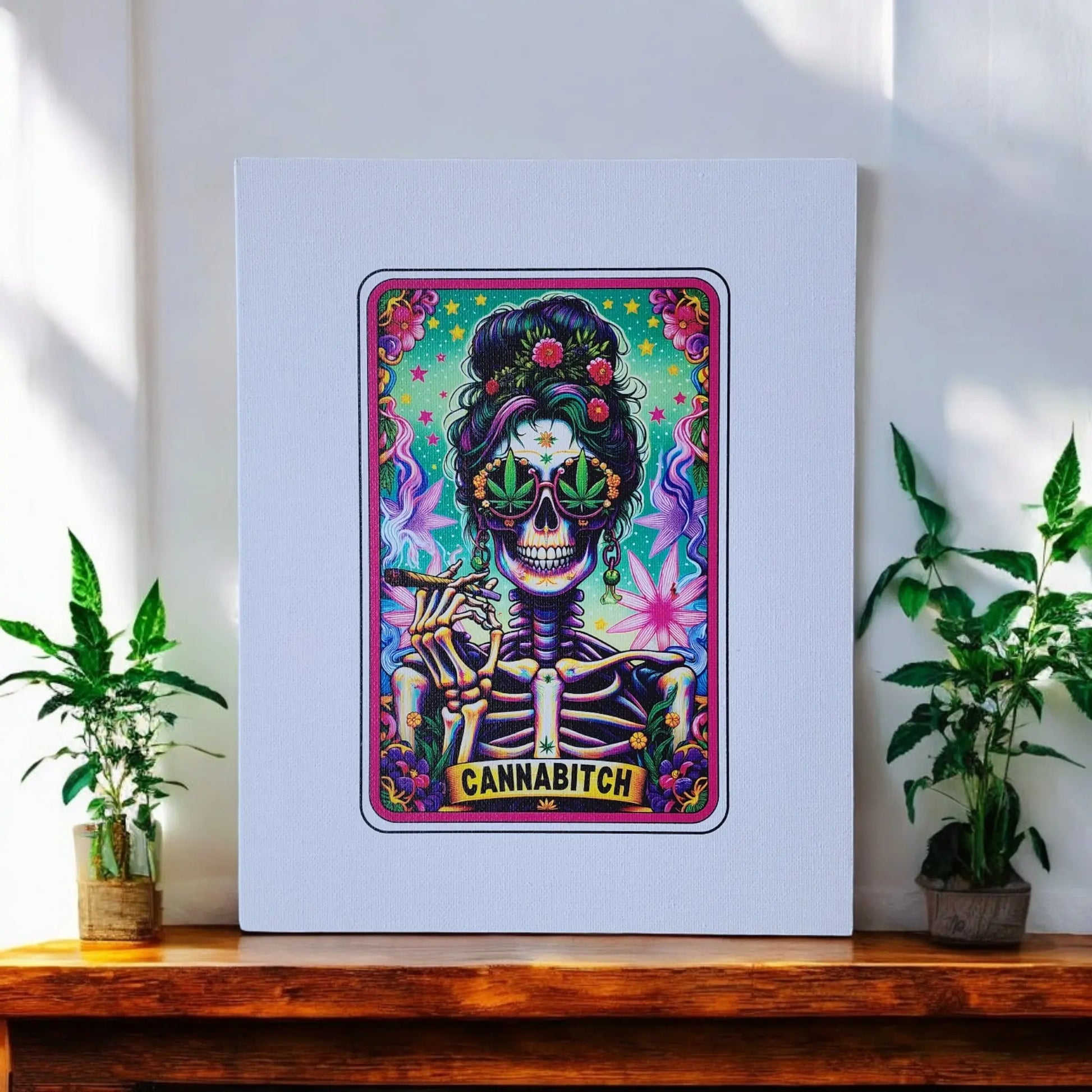 A vibrant, psychedelic-style painting resembling a tarot card design features a skeleton adorned with a floral crown and colorful sunglasses, holding a cigarette, with the word "CANNABITCH" inscribed at the bottom. This distinctive Cannabitch 8x10 Canvas Sign by Mirabilia Boutique is displayed on a wooden shelf alongside potted plants against a white wall.