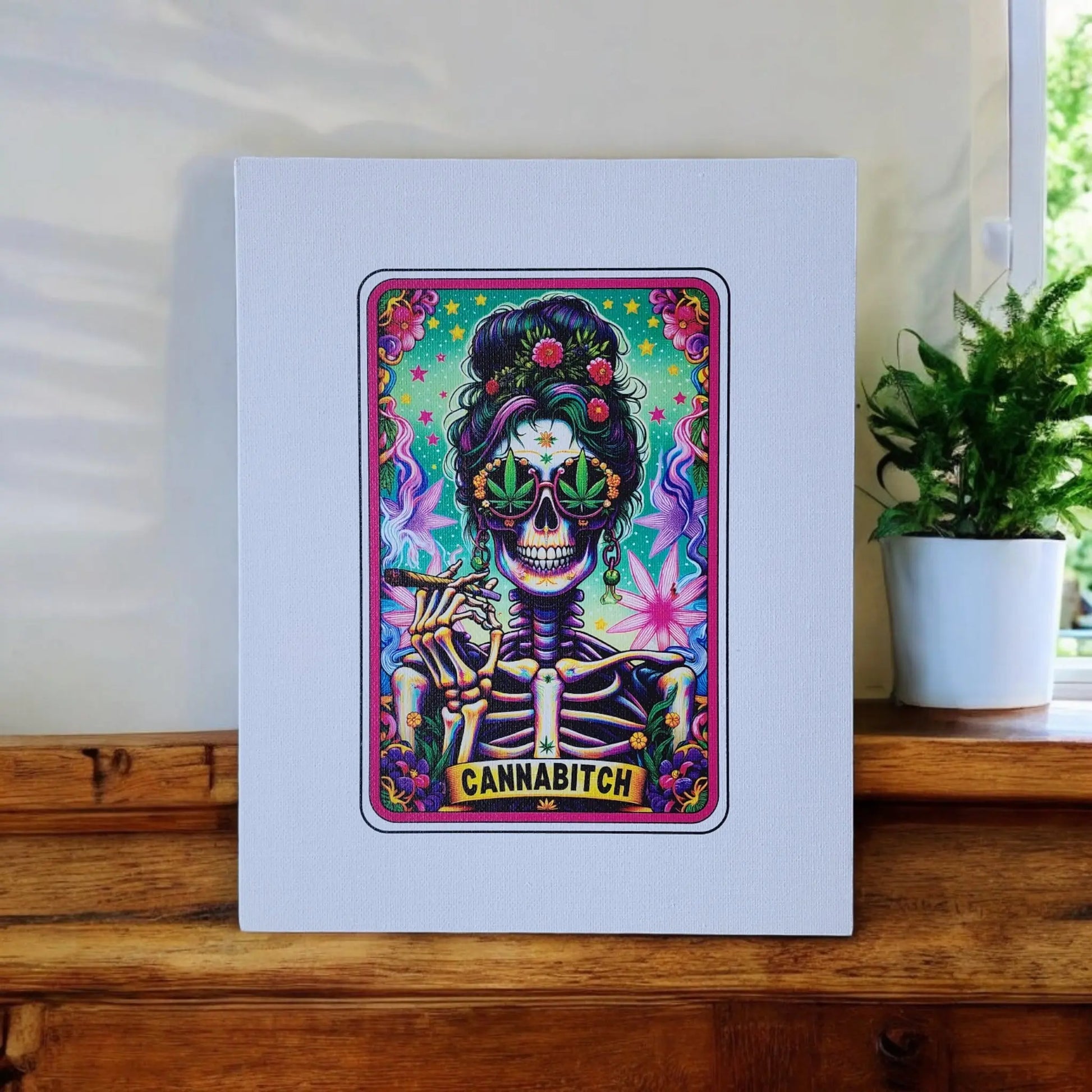 The Cannabitch 8x10 Canvas Sign by Mirabilia Boutique showcases a vibrant art print titled "Cannabitch," featuring a skeleton with sunglasses, adorned with colorful flowers and cannabis leaves. Embracing cannabis culture art, the skeleton holds a marijuana joint. The Cannabitch Canvas Sign is displayed on a wooden surface beside a small potted plant.