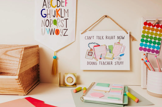 A teacher's desk features colorful supplies, including notebooks, pens, and sticky notes. Classroom decor incorporates a wall hanging with the alphabet and a "Can't Talk Right Now: Teacher Stuff" sign by Mirabilia Boutique. A wooden organizer displays vibrant push pins.