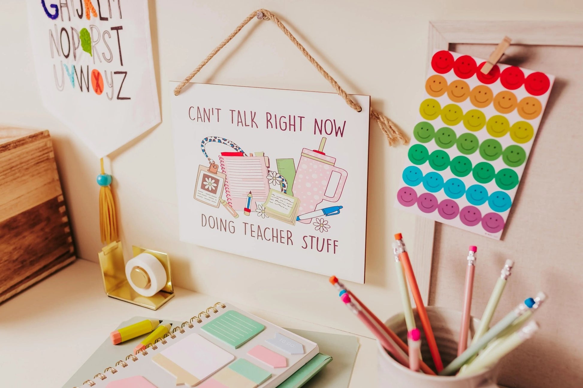 A cozy desktop features colorful sticky notes, pencils in a jar, and a whimsical wall sign from Mirabilia Boutique called the "Can't Talk Right Now: Teacher Stuff Hanging Sign." A rainbow of smiley face stickers adds charm, while vibrant wall art showcases alphabet letters as delightful classroom decor.