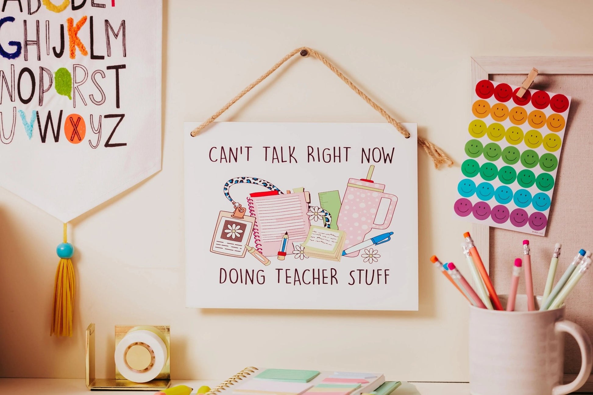 A "Can't Talk Right Now: Teacher Stuff Hanging Sign" by Mirabilia Boutique adorns the wall, featuring the phrase and surrounded by school-themed illustrations. This delightful classroom decoration is complemented by vibrant cards, pencils in a cup, and an alphabet banner that enlivens the area.