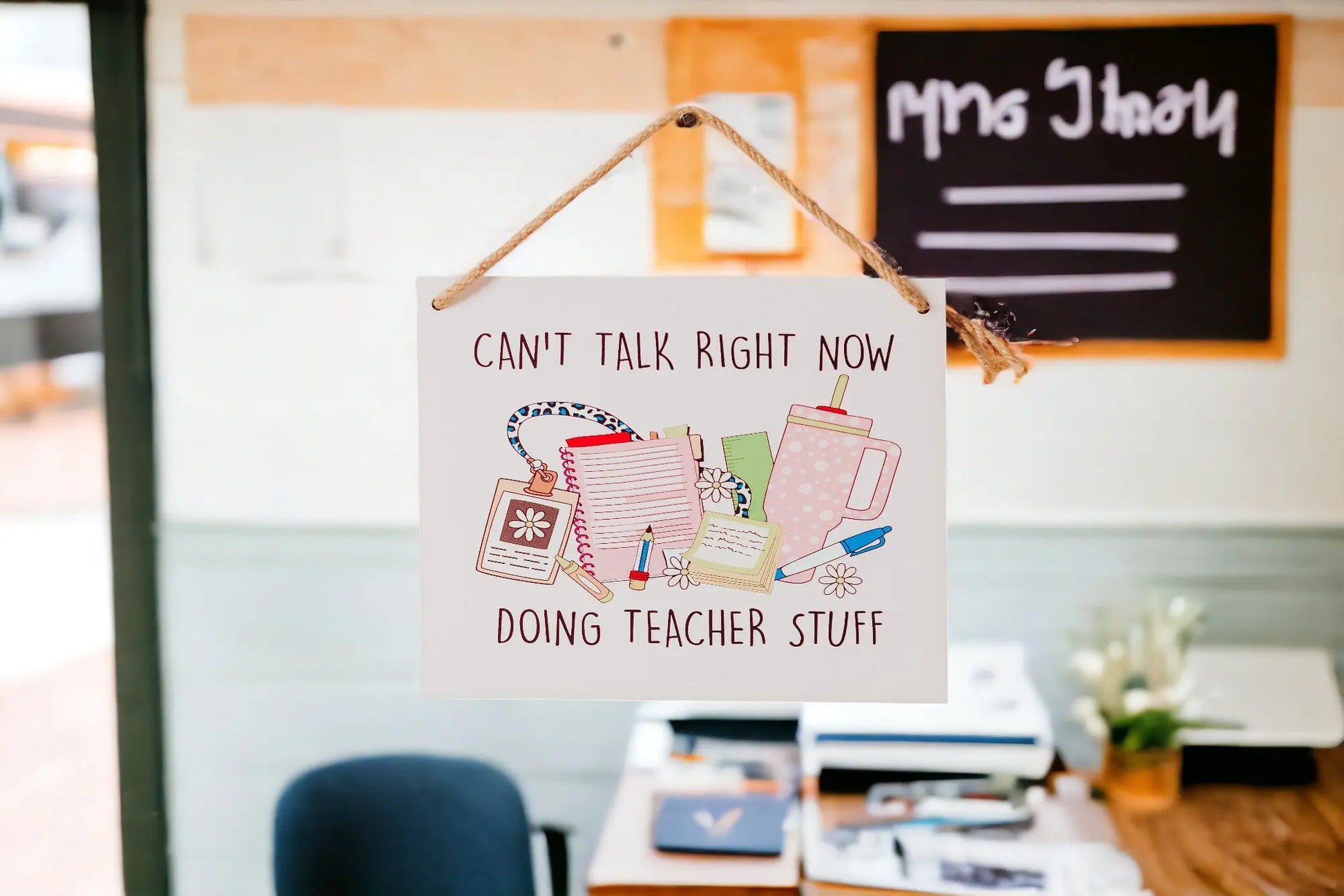 The "Can't Talk Right Now: Teacher Stuff Hanging Sign" by Mirabilia Boutique is a delightful classroom decor piece that displays the phrase "Can't talk right now, doing teacher stuff." It is adorned with illustrations of a notepad, marker, apple, coffee cup, and paper clips. In the background, there’s a desk with papers and a chair. This charming sign is perfect for adding personality to any teaching space.