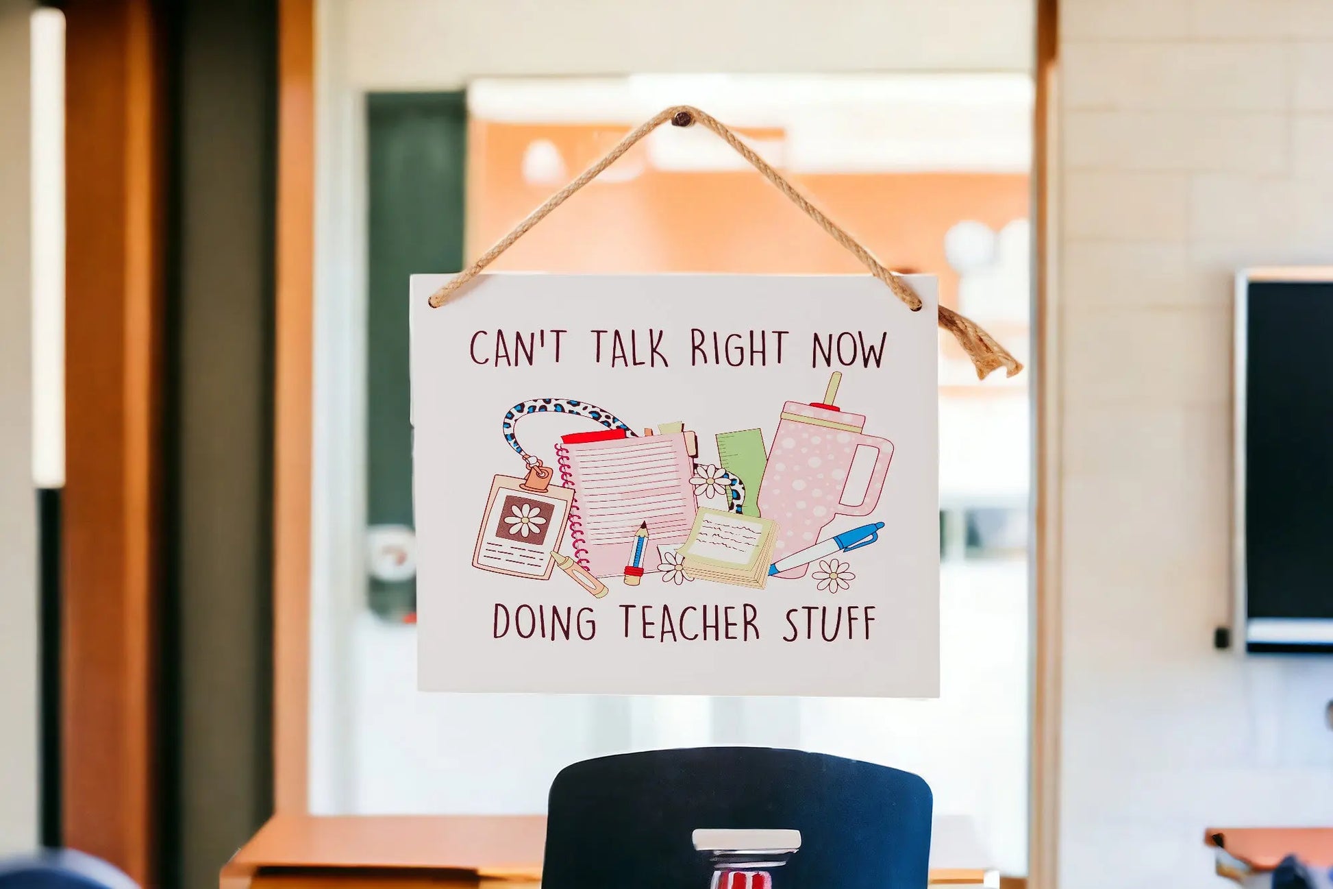 A wall sign from Mirabilia Boutique, named "Can't Talk Right Now: Teacher Stuff Hanging Sign," drapes over a chair, displaying the message, "Can't talk right now, doing teacher stuff." It features illustrations of books, a pen, coffee tumbler, and notepad against a backdrop of a blurred classroom decor setting.