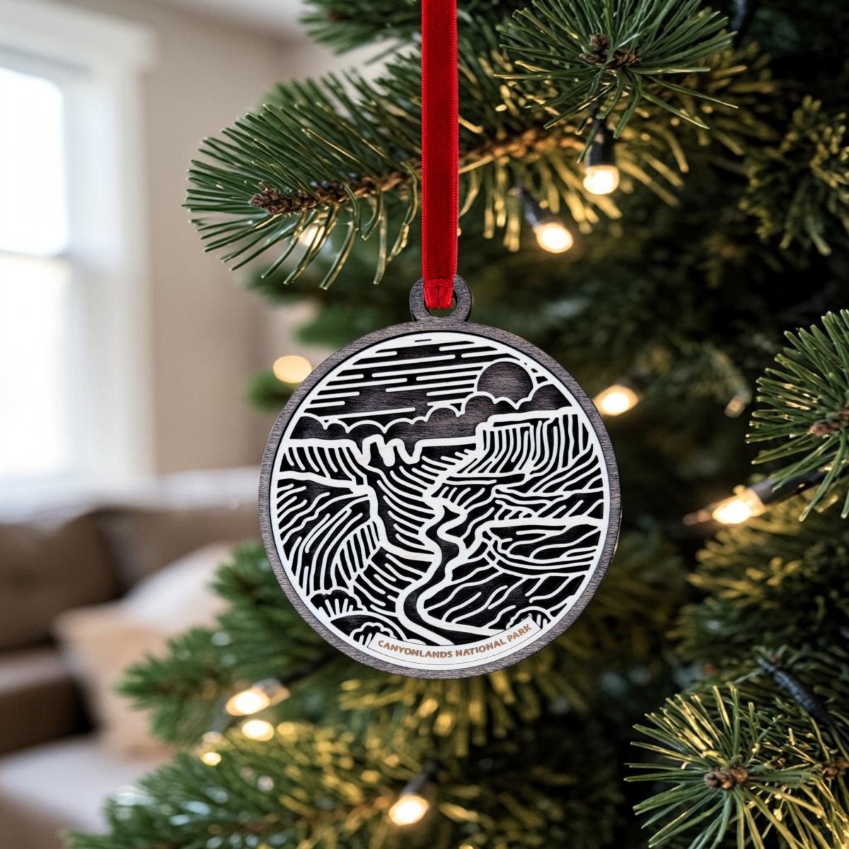 A round, black and white wooden Canyonlands National Park Christmas Ornament by Mirabilia Boutique, featuring intricate line art of a scenic landscape, hangs from a red ribbon on a Christmas tree adorned with white lights. This handcrafted keepsake adds charm to the festive scene, while a blurred couch and window provide a cozy background.