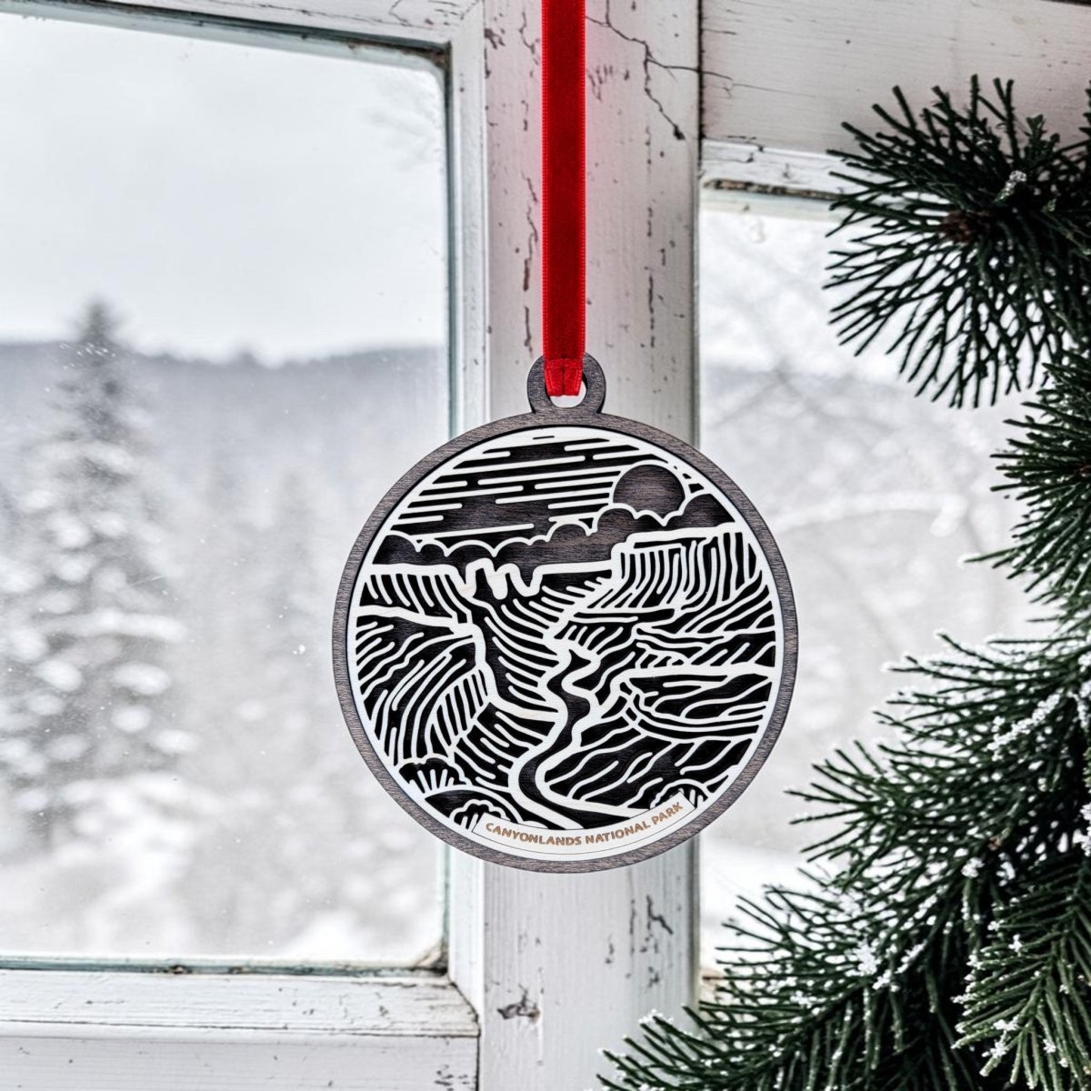 A Canyonlands National Park Christmas Ornament from Mirabilia Boutique, featuring an oval black and white landscape design, hangs by a red ribbon near a frosty window with snowy trees and pine branches visible outside.