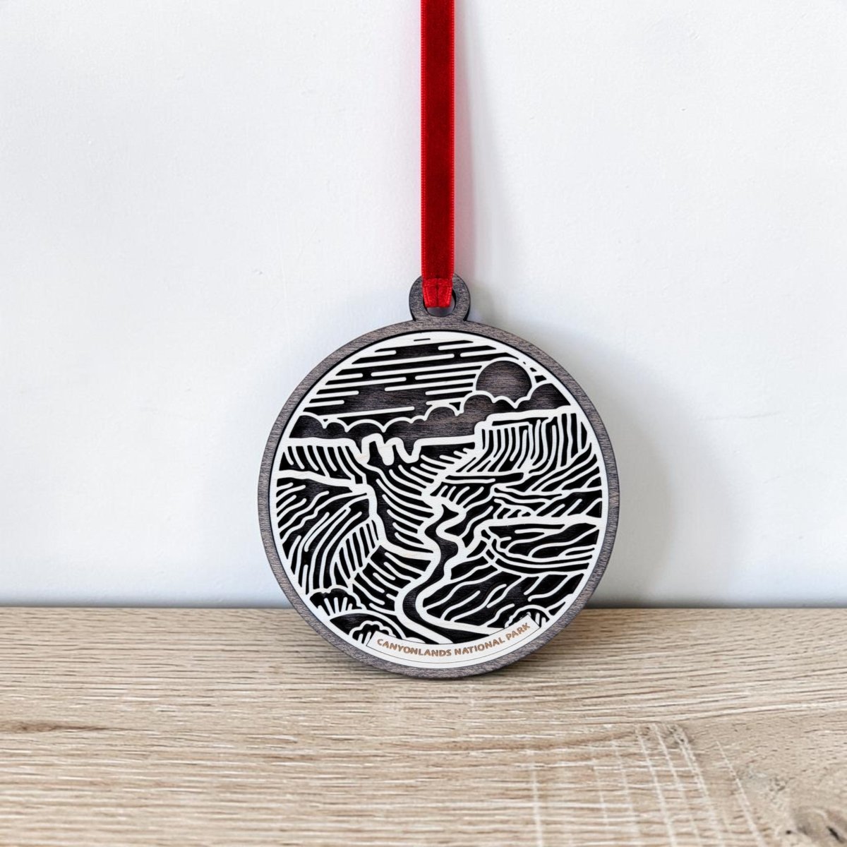 The Canyonlands National Park Christmas Ornament by Mirabilia Boutique is a round, handcrafted keepsake featuring a black and white landscape design with hills, a river, and the sun. It hangs from a red ribbon against a white wall and rests on a light wooden surface.
