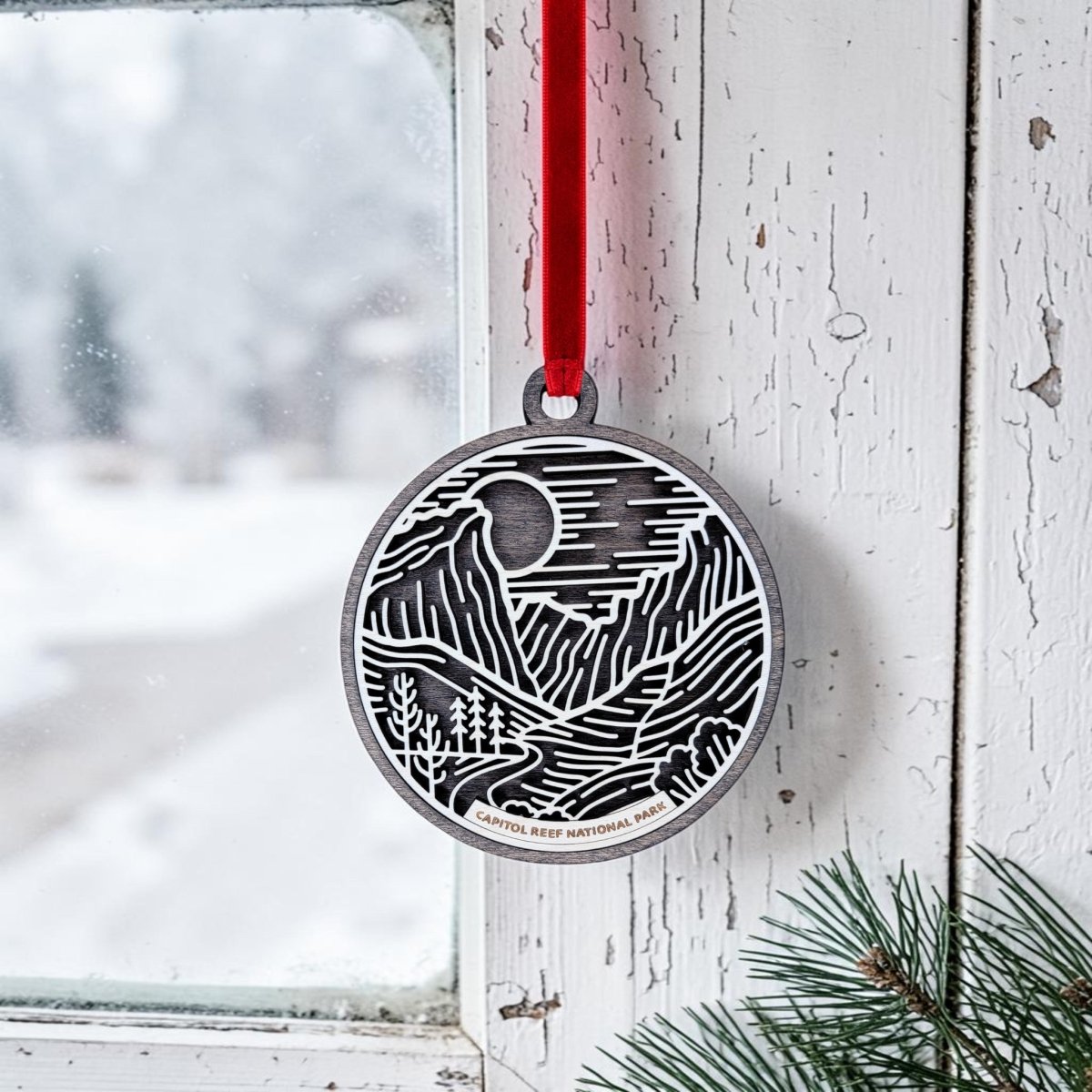 A round metal Capitol Reef National Park Line Art Ornament by Mirabilia Boutique, featuring a mountain and tree design, hangs by a red ribbon on a snowy window. Nearby, pine needles rest on the sill. The ornament is labeled "CAPITOL REEF NATIONAL PARK.