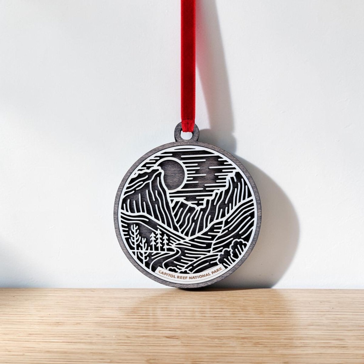 The Capitol Reef National Park Line Art Ornament by Mirabilia Boutique is a round wooden ornament featuring an engraved landscape of mountains, trees, and either a sun or moon. It hangs from a red ribbon and includes a label at the bottom that reads "Capitol Reef National Park." The ornament is displayed on a light wooden surface.