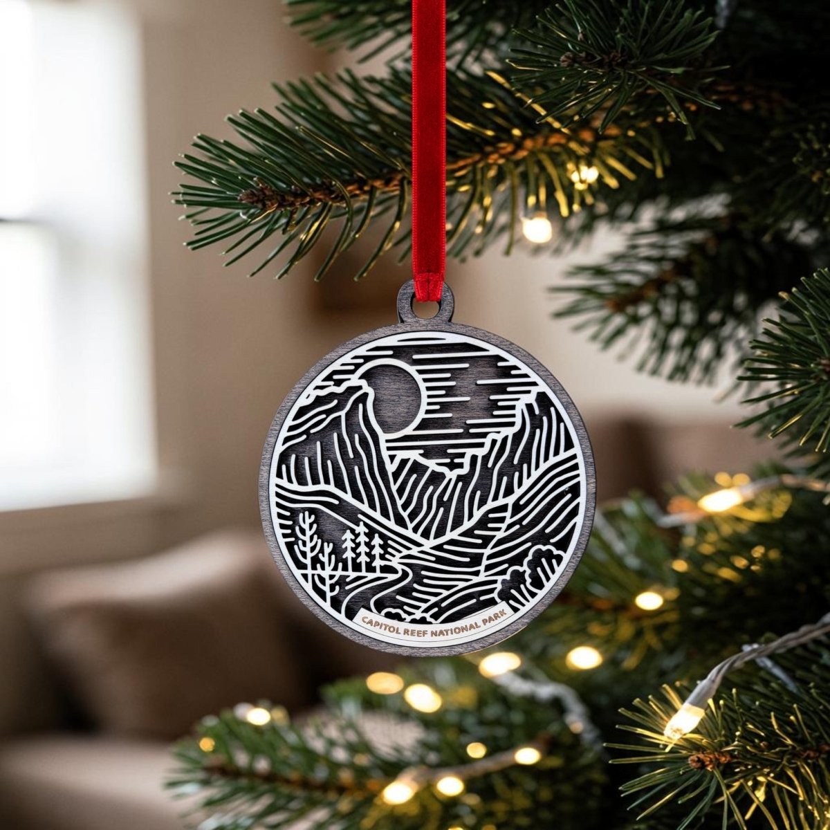 A silver Capitol Reef National Park Line Art Ornament by Mirabilia Boutique, featuring a detailed landscape design, hangs from a red ribbon on a Christmas tree branch. Warm string lights illuminate the ornament, which reads "Capitol Reef National Park," set against a softly blurred indoor background.