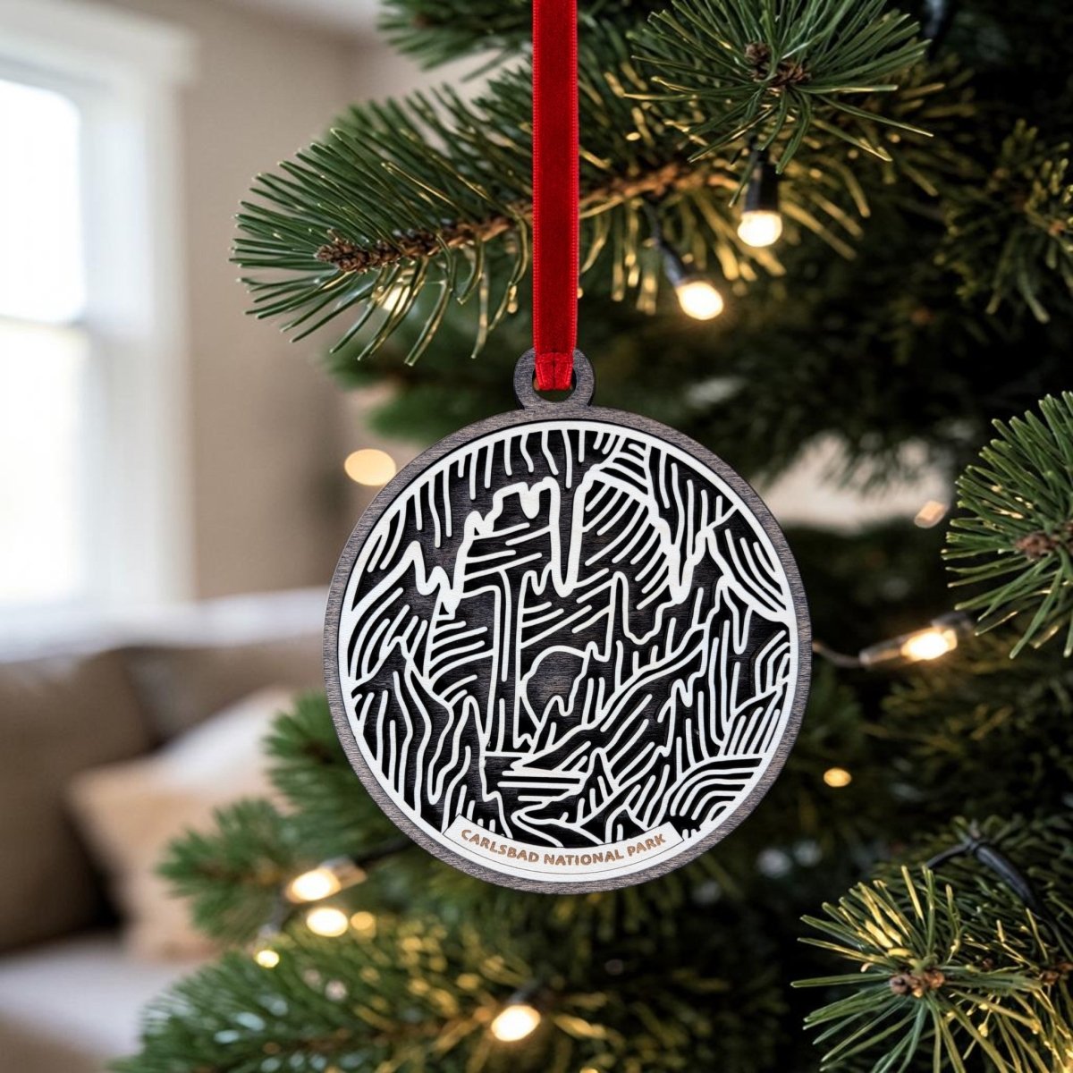 A Carlsbad Caverns National Park Christmas Ornament from Mirabilia Boutique, featuring a striking black and white cave design, hangs from a red ribbon on the Christmas tree. The handcrafted wooden ornament is inscribed with “Carlsbad National Park,” celebrating the wonder of Carlsbad Caverns. Warm lights and a blurred couch in the background enhance the cozy holiday atmosphere.