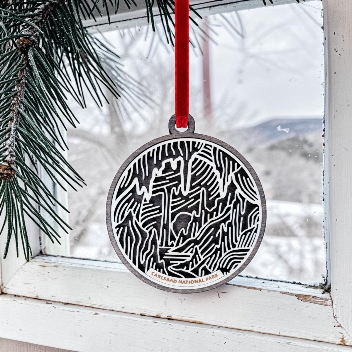 A round, handcrafted wooden ornament from Mirabilia Boutique—featuring a cave design and the words "Carlsbad National Park"—hangs by a red ribbon on a windowpane. Pine branches and a snowy landscape outside set the scene, celebrating the beauty of Carlsbad Caverns National Park with this festive Carlsbad Caverns National Park Christmas Ornament.
