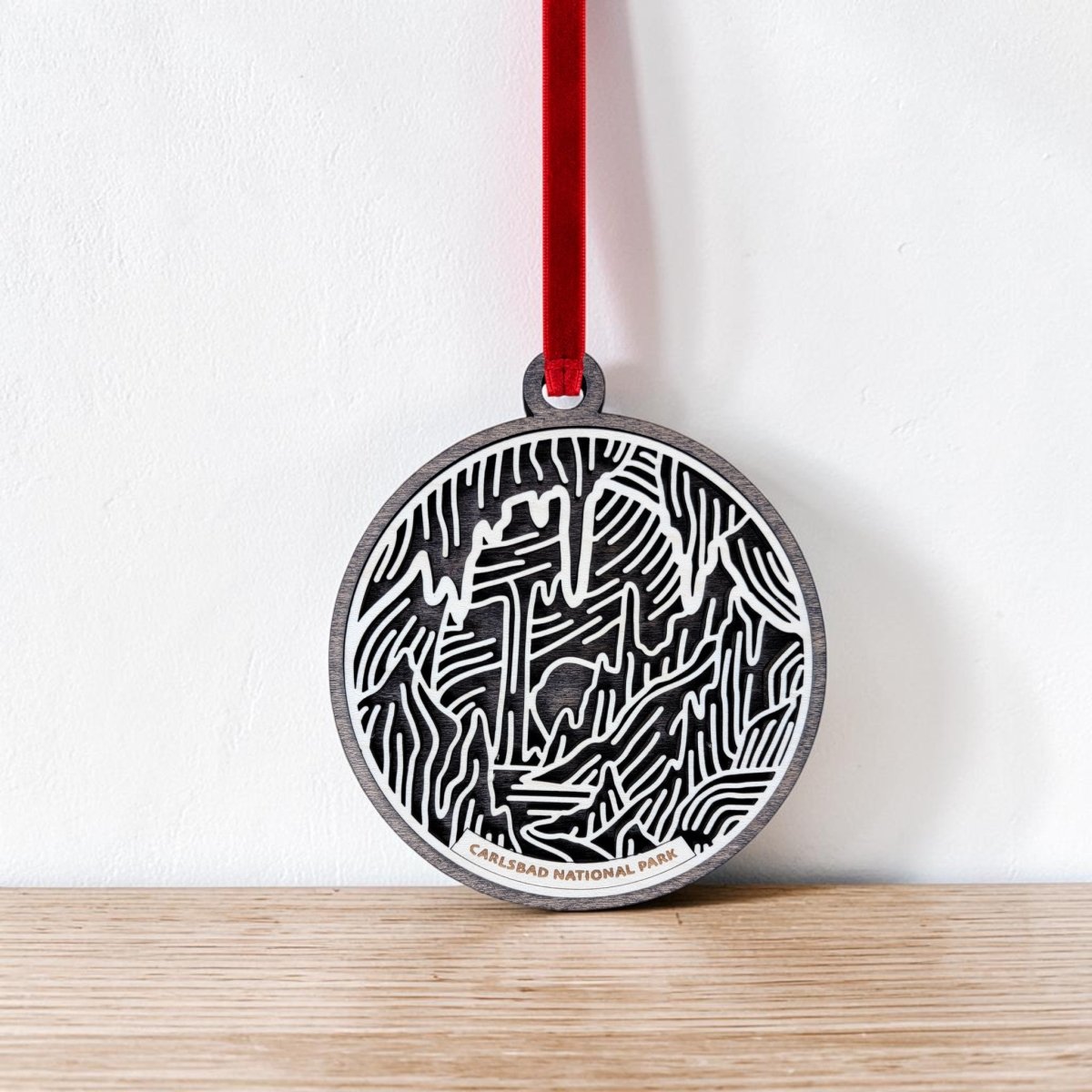 Crafted by Mirabilia Boutique, the Carlsbad Caverns National Park Christmas Ornament is a beautifully handcrafted wooden piece featuring detailed black and white cave-inspired designs. It hangs gracefully from a red ribbon, and "Carlsbad Caverns National Park" is engraved at the bottom—making it an ideal ornament for collectors and nature lovers.