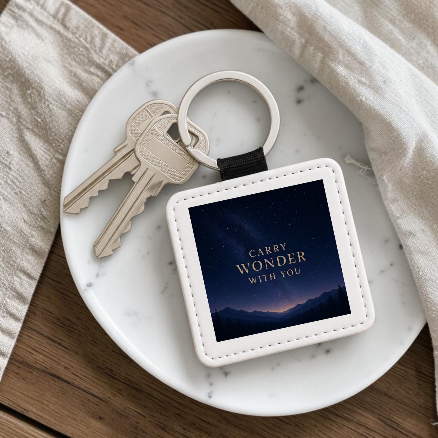 The Carry Wonder Celestial Keychain from Mirabilia Boutique’s Star & Solstice Collection rests on a white plate with two keys attached. This celestial accessory features a night sky, mountain silhouette, and the phrase “CARRY WONDER WITH YOU” in gold text. A cream-colored cloth lies nearby, highlighting this decorative keepsake’s charm.